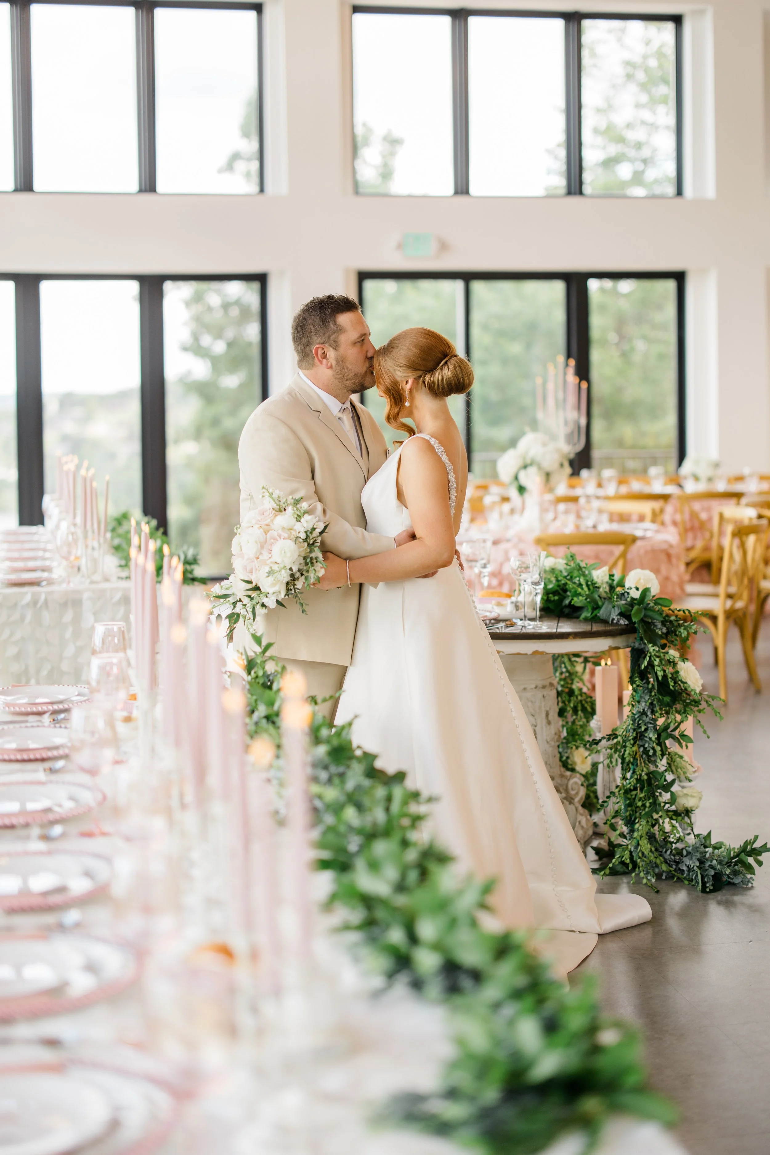 A bride and groom kissing at their wedding reception, with decorated tables and large windows in the background.