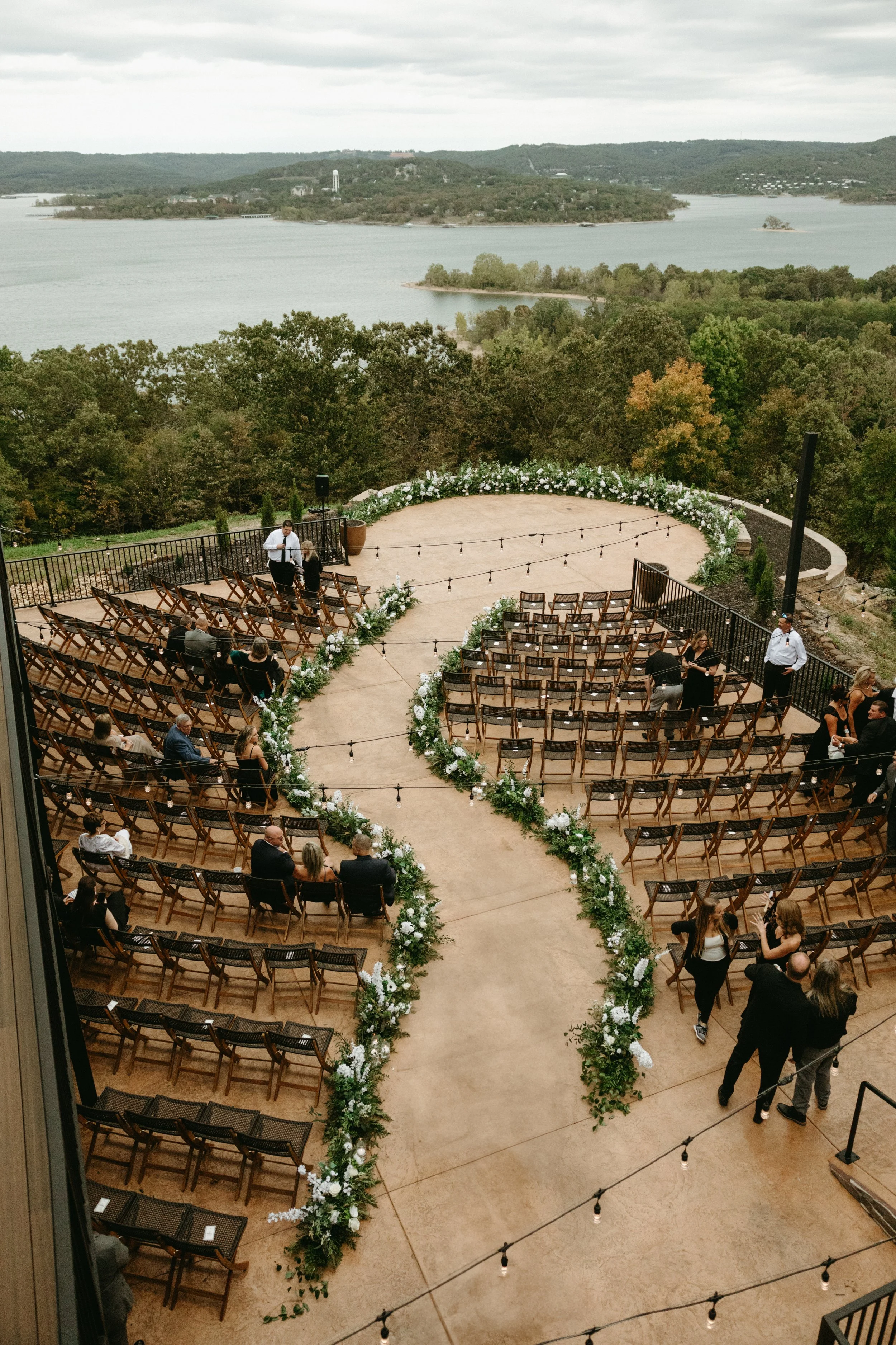 Outdoor wedding ceremony setup with chairs arranged in rows on either side of a floral aisle, overlooking a lake and wooded landscape under an overcast sky in the Ozarks.