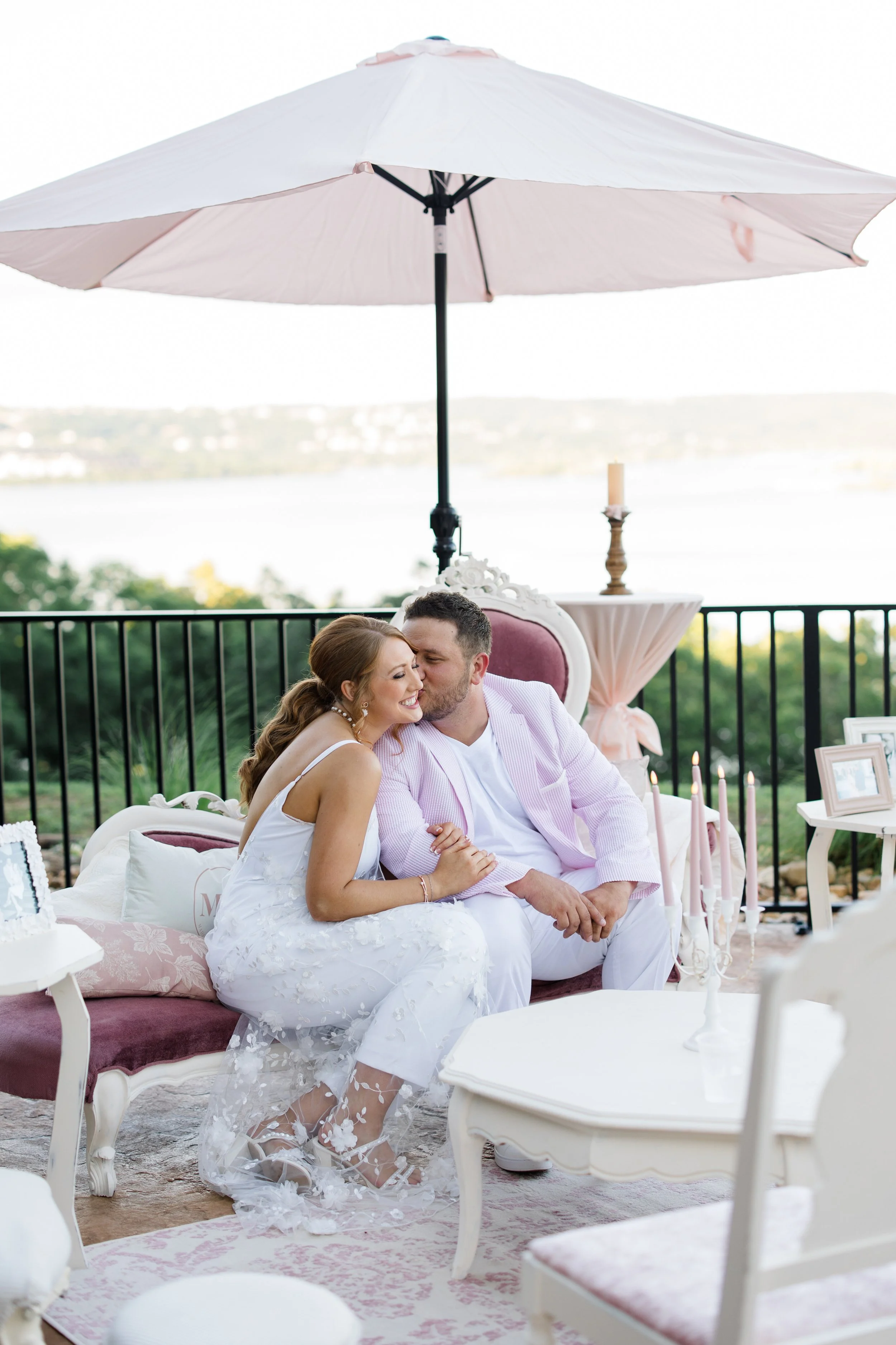 A couple sitting on a vintage loveseat under a pink and white patio umbrella, with a scenic outdoor lake view in the background. The woman is smiling and leaning into the man, who is kissing her cheek.
