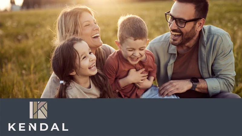 A family sitting outdoors on grass, smiling and laughing together during sunset, with two children and two adults.