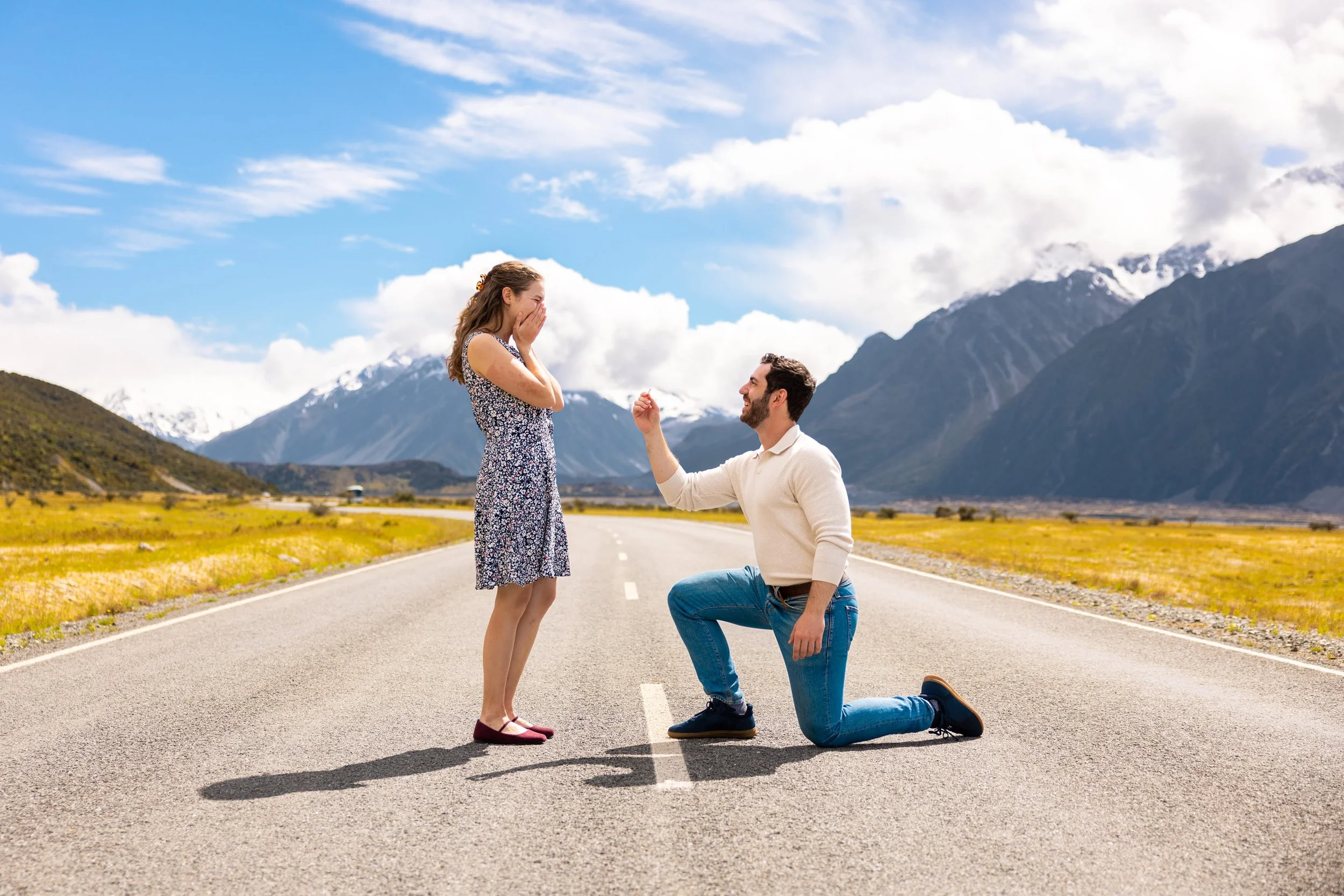 A man proposing to a woman on a road with mountains and clouds in the background.