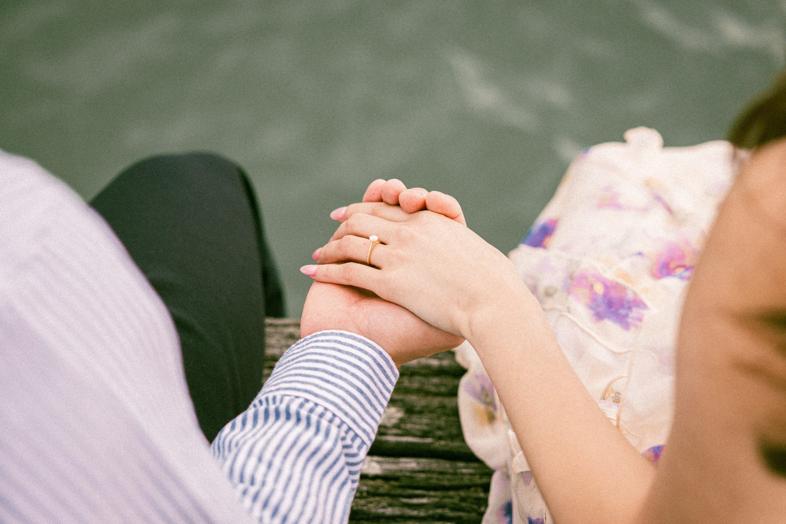 Close-up of a man and woman holding hands, sitting by the water on a wooden dock, with the woman's engagement ring visible.