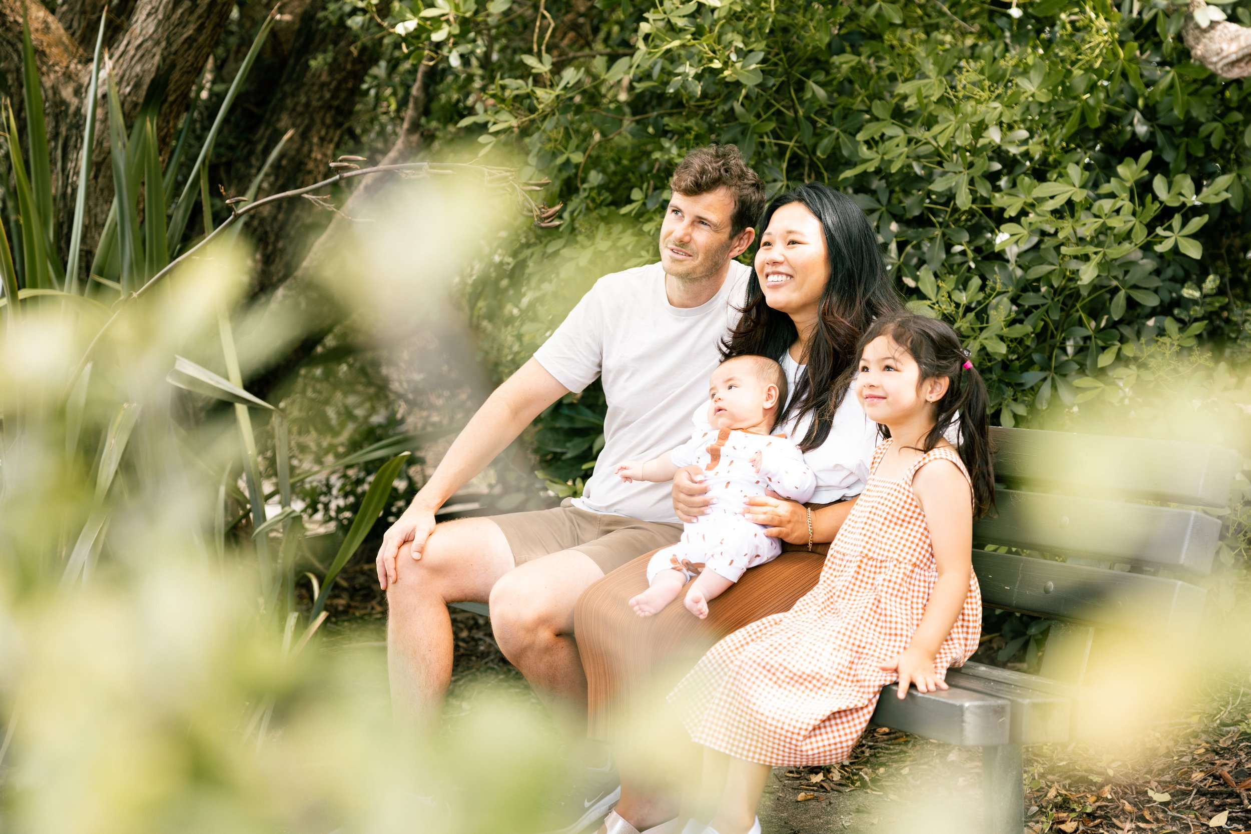 A family of four sitting on a park bench surrounded by green foliage. The father, mother, and two young daughters are smiling and enjoying a moment together.