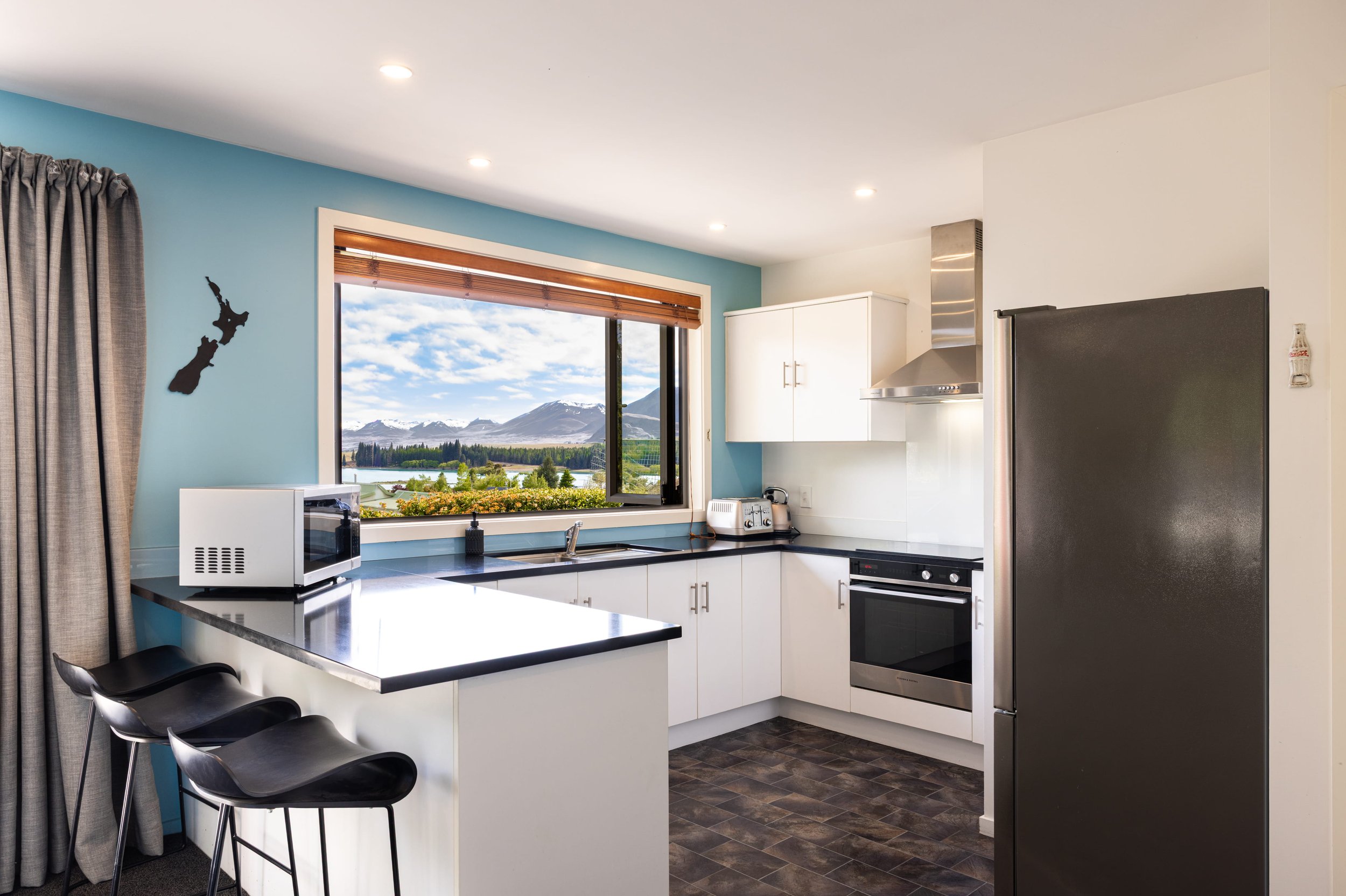 Modern kitchen with white cabinets, black countertop, stainless steel oven and range hood, black refrigerator, microwave, toaster, and a window with mountains in the background.