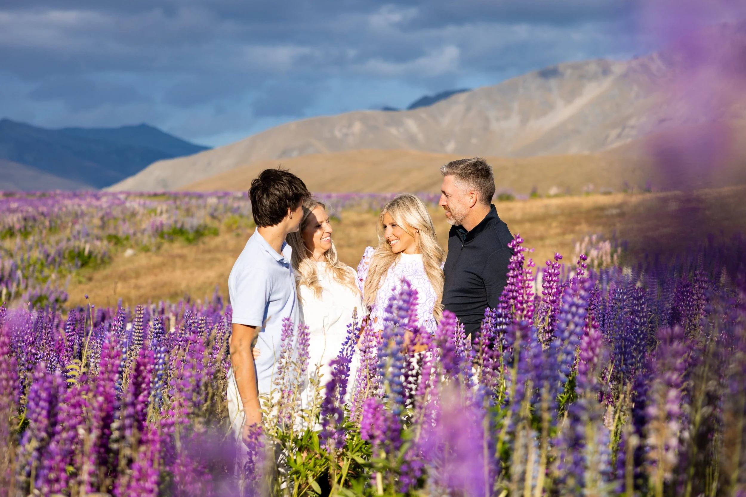 Four people standing in a field of purple flowers with mountains in the background, smiling and looking at each other.