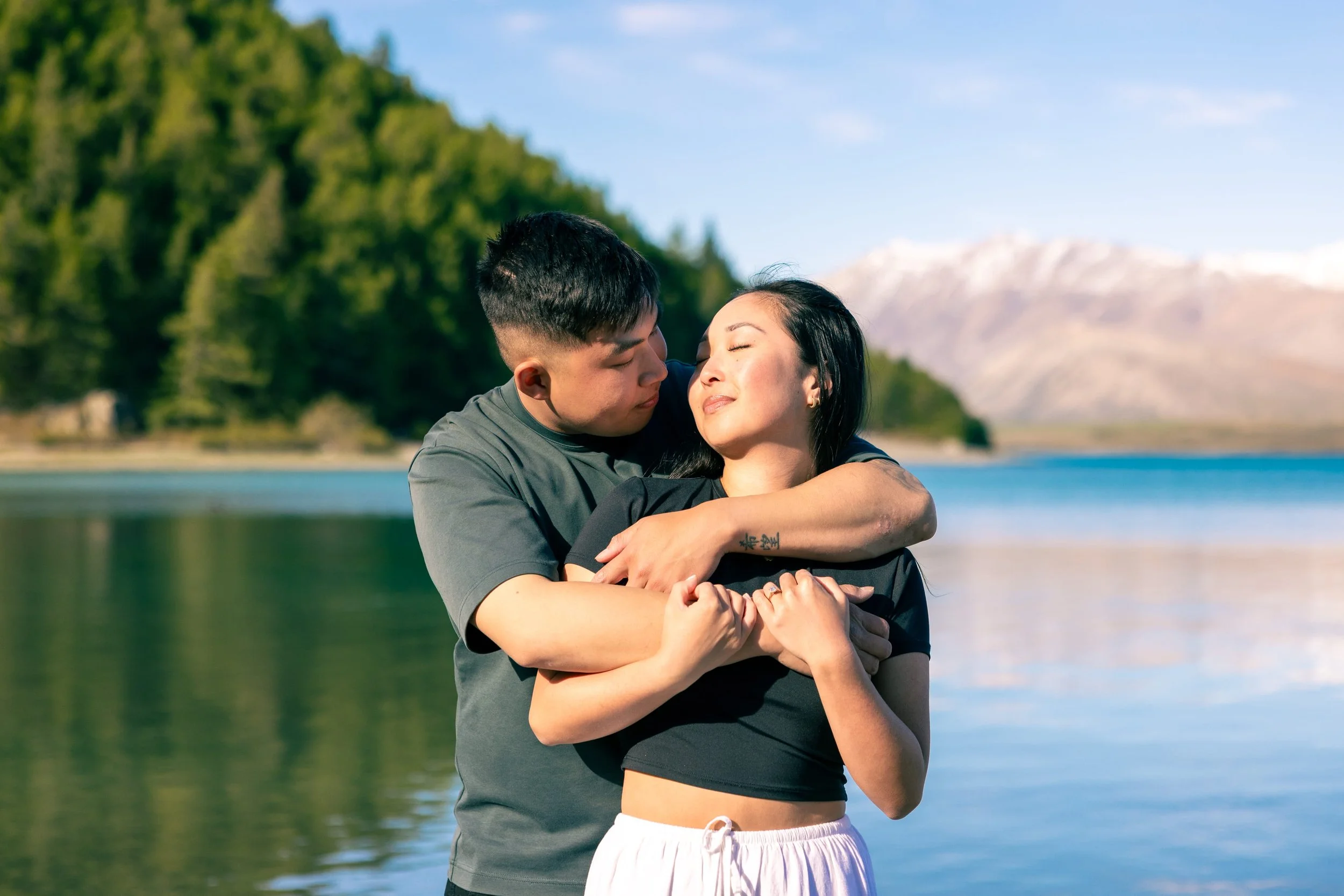 A couple embracing outdoors near a lake with mountains in the background, surrounded by trees, on a sunny day.