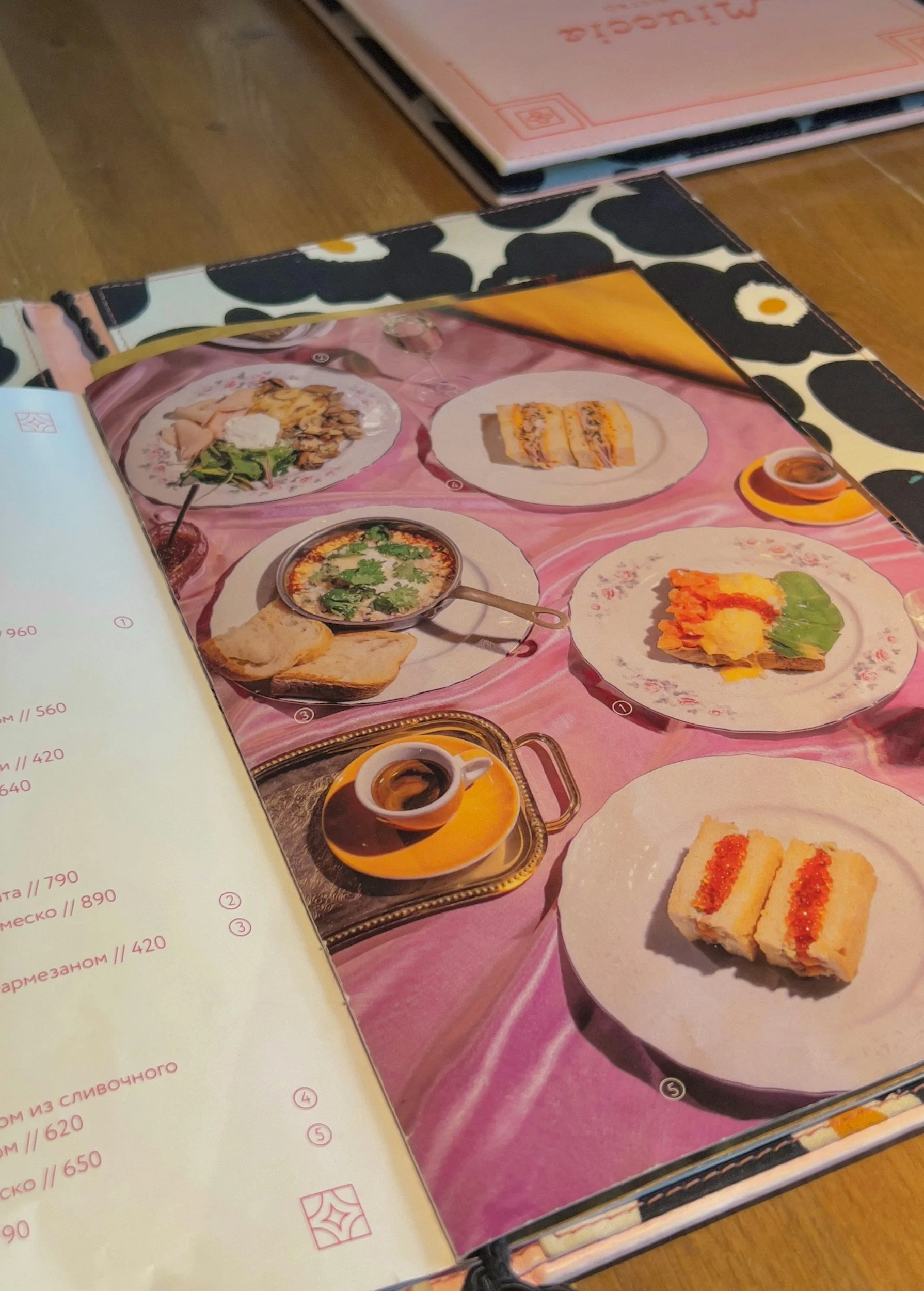 A menu page displaying various dishes, including rolled sandwiches, a bowl of soup, and small sandwiches, set on a pink tablecloth with a black and white patterned border and glasses of water.