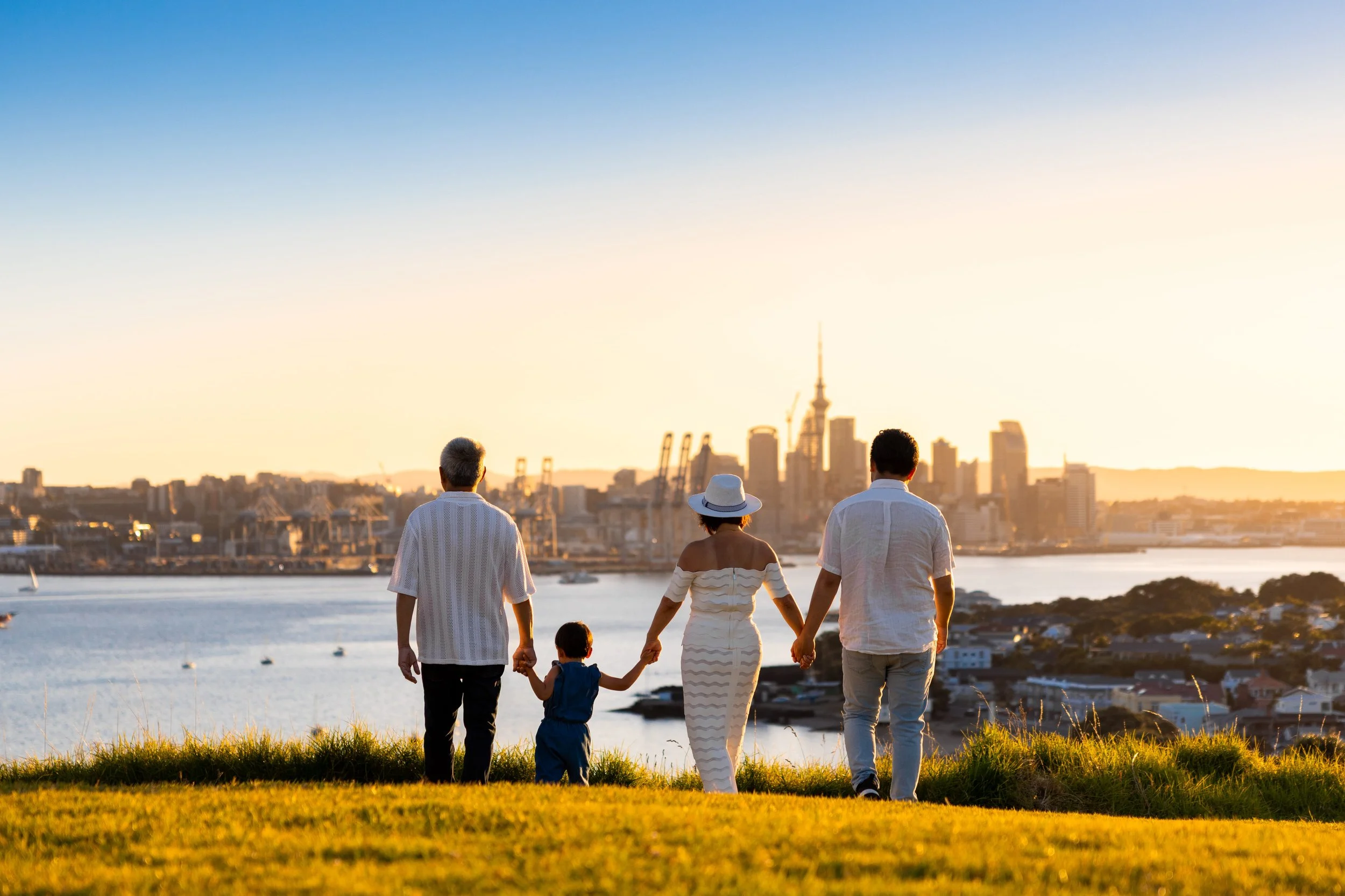Family of five walking hand in hand on a grassy hill at sunset with a city skyline and water in the background.