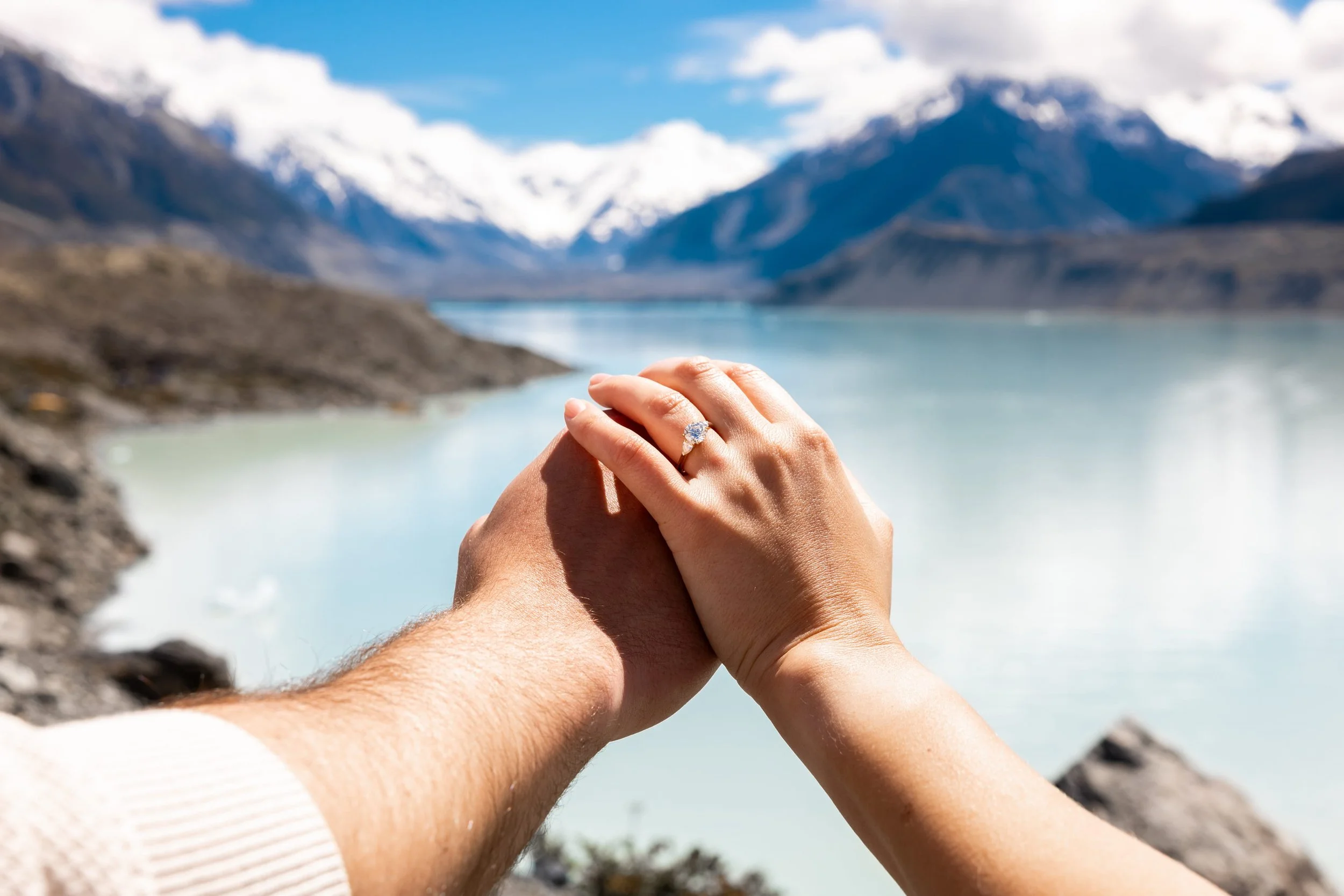 A couple holding hands with an engagement ring in front of a mountain lake with snow-capped mountains in the background.