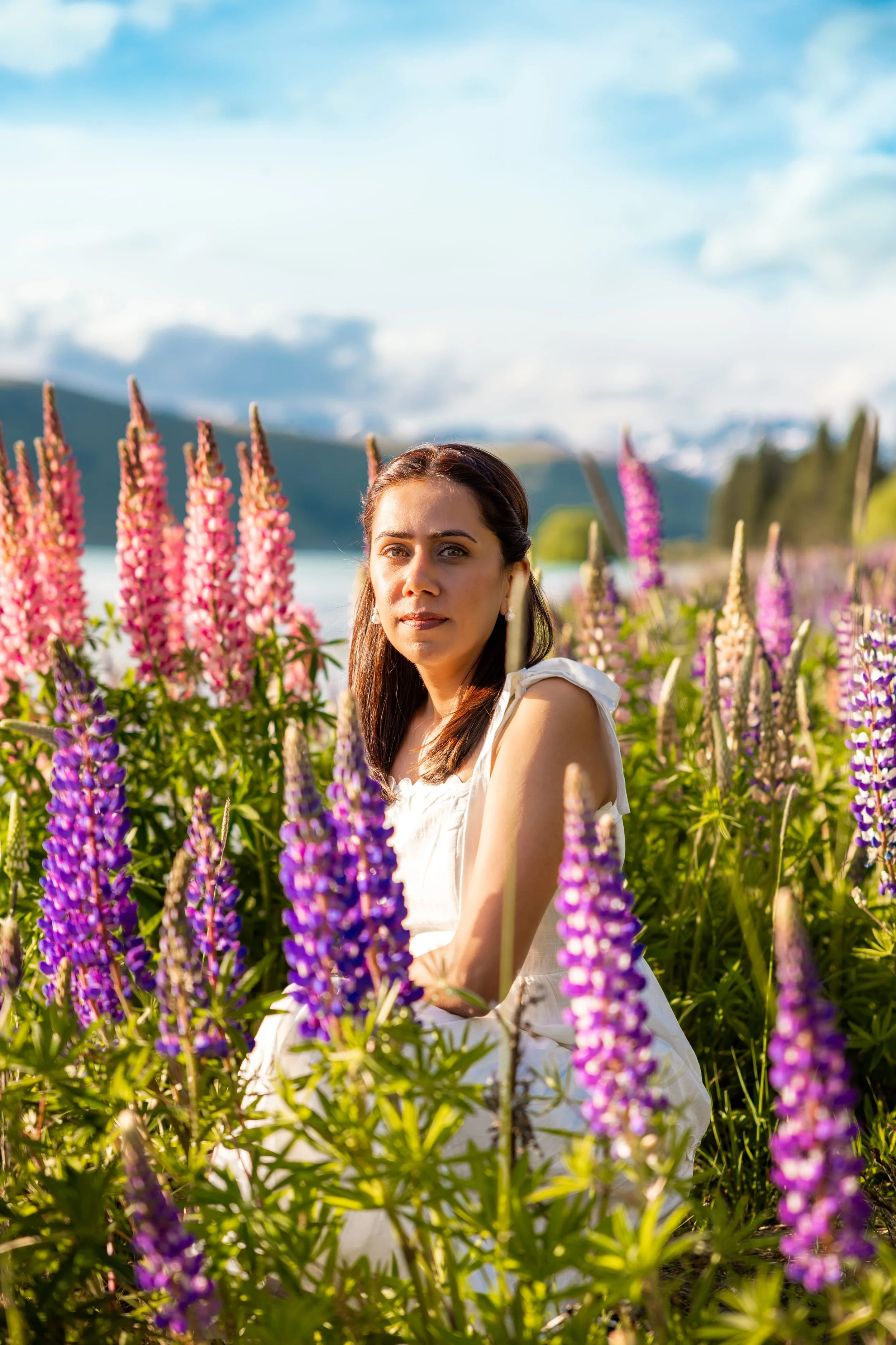 A woman in a white dress sitting among pink and purple lupine flowers outdoors with a lake, hills, and a blue sky in the background.