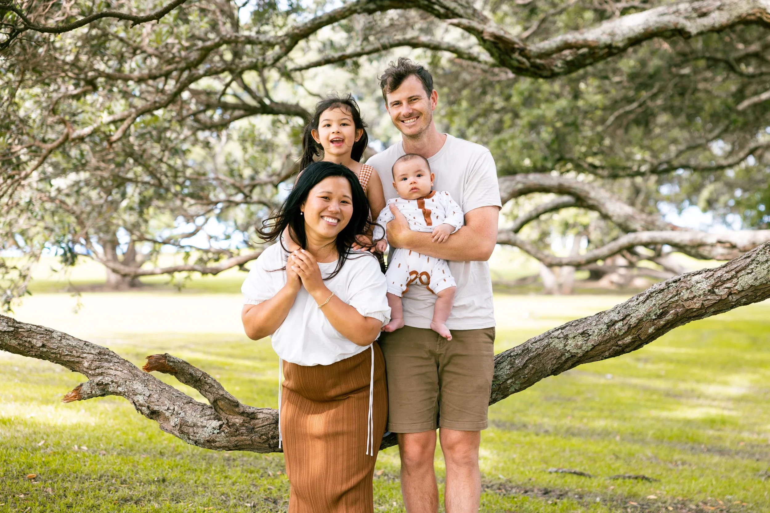 A family of four with two children standing on a tree branch outdoors in a park.