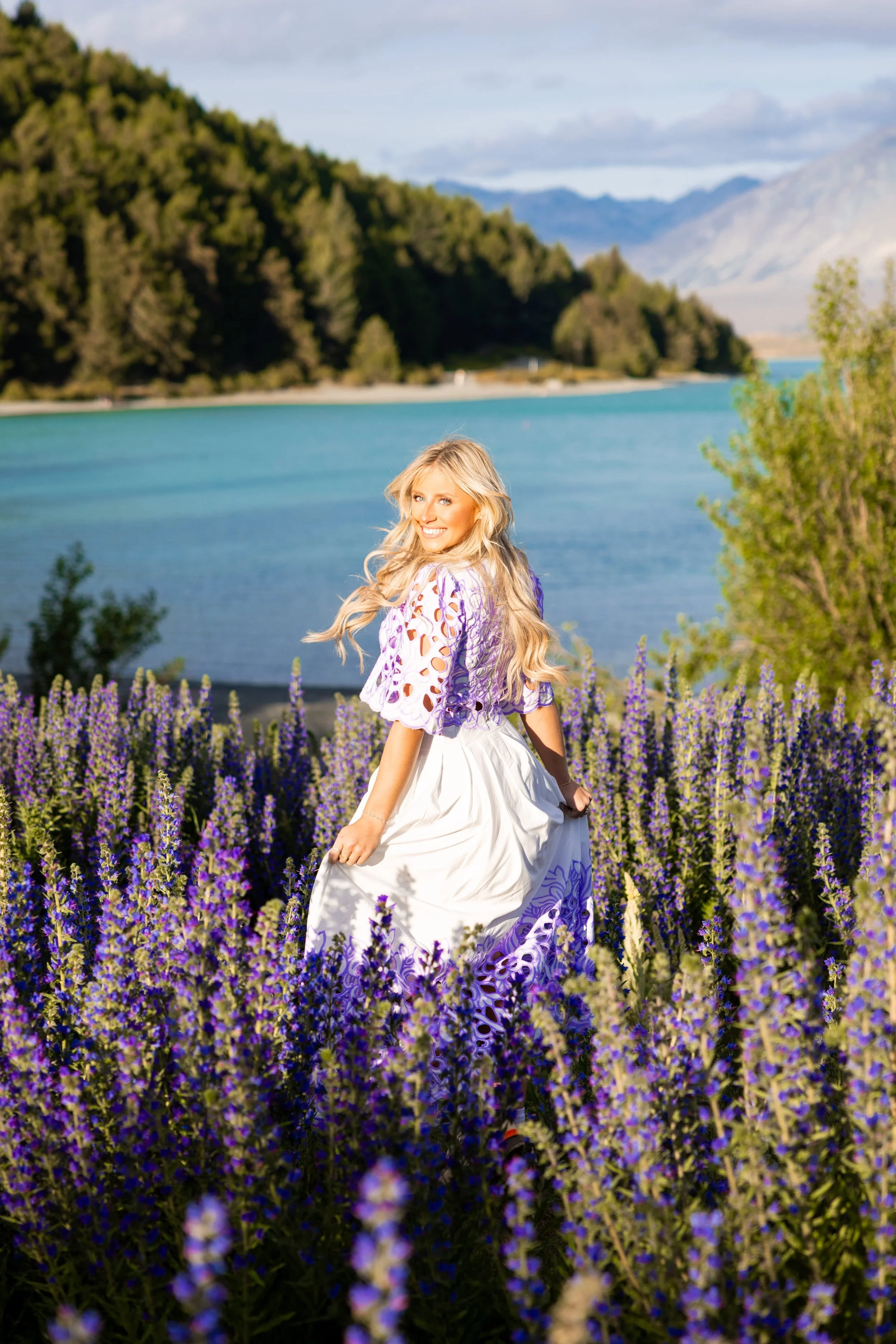 A woman in a white and purple dress standing in a field of purple flowers near a lake, with mountains in the background and a partly cloudy sky.