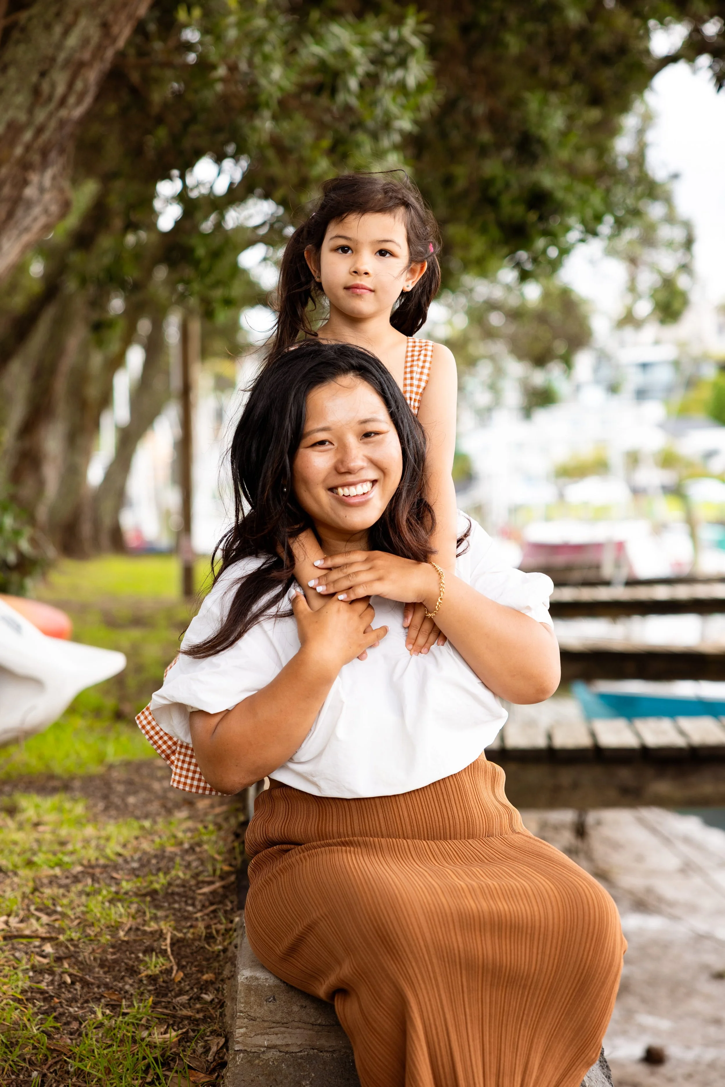 A woman and a young girl sitting outdoors near a tree, with boats and water in the background. The girl is on the woman's shoulders, and they are smiling and posing for the camera.