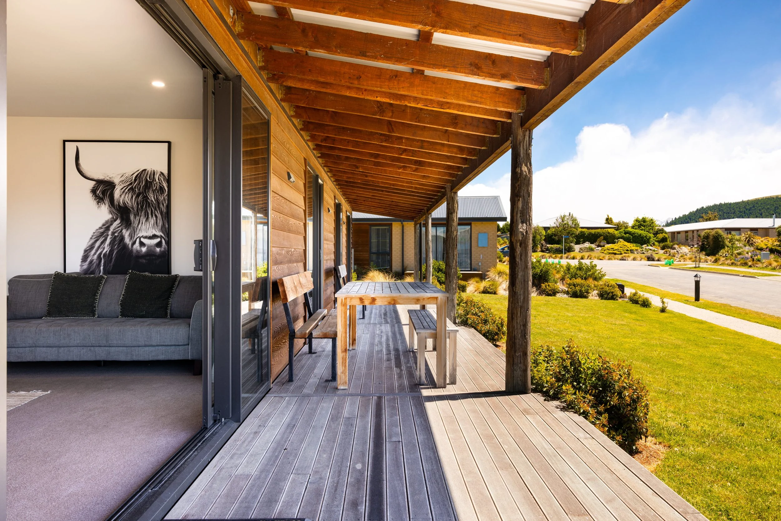 View of a wooden house patio with a dining table and benches, overlooking a garden and neighboring houses on a sunny day.