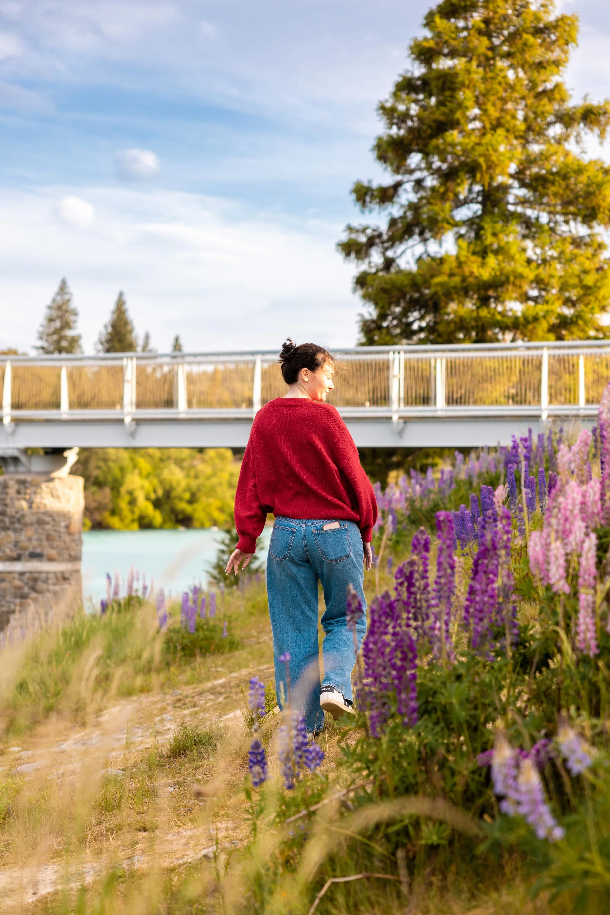 A woman in a red sweater and blue jeans walking along a path beside purple and pink flowers during daytime, with a bridge and trees in the background.
