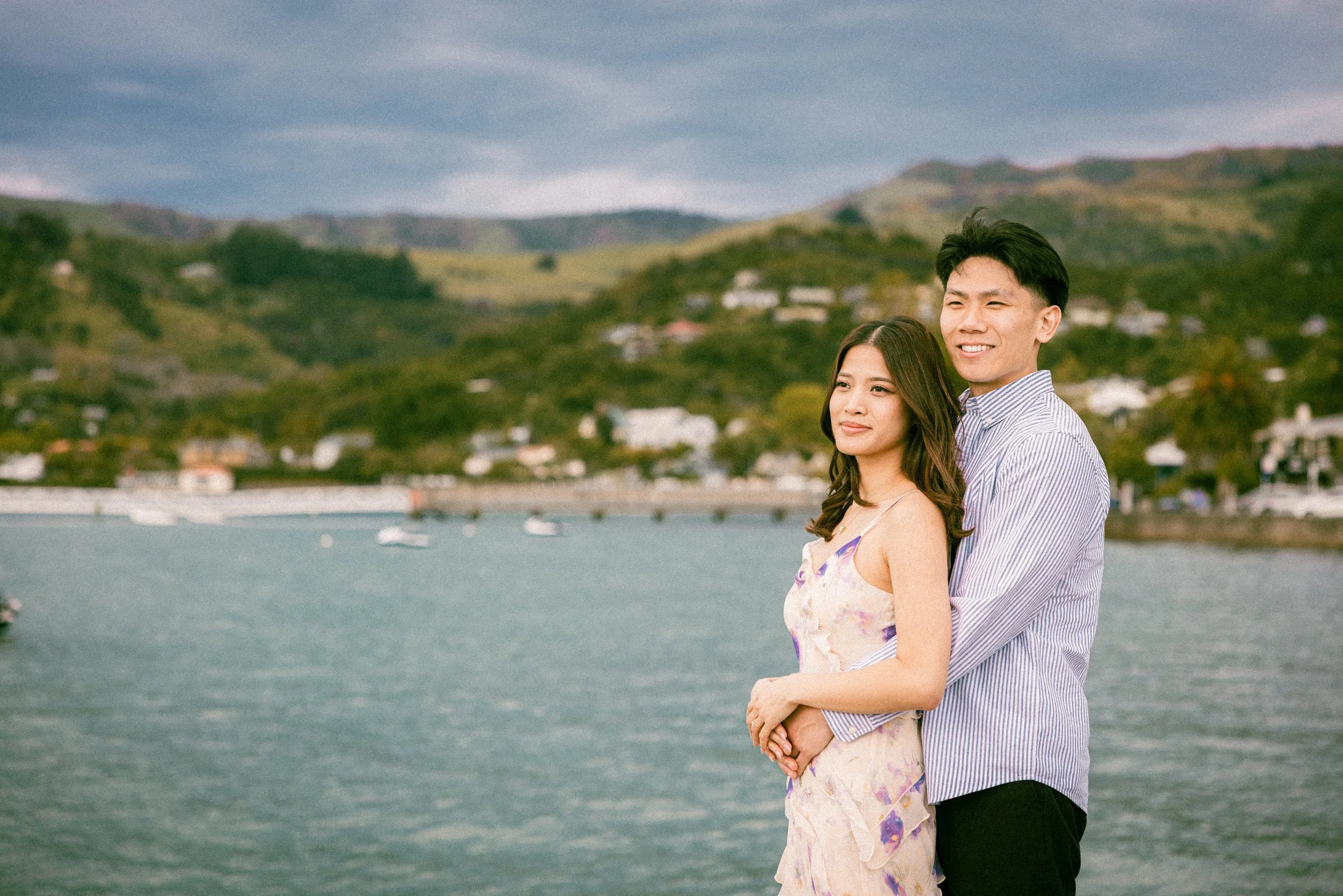 A young couple standing by a body of water with boats, green hills, and houses in the background on a cloudy day.