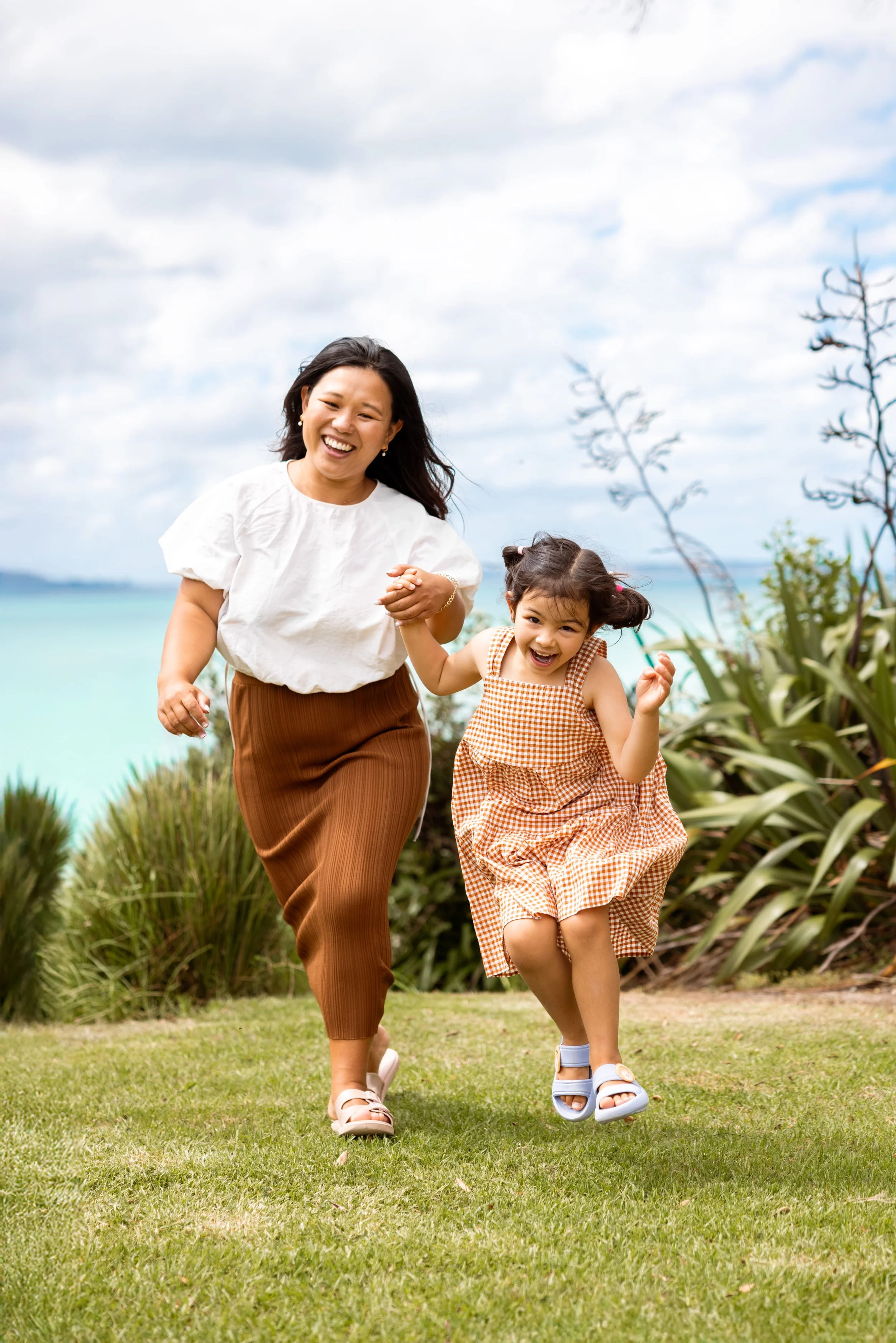 A woman and a young girl are running hand in hand outdoors near a body of water, with smiles and joyful expressions on their faces.
