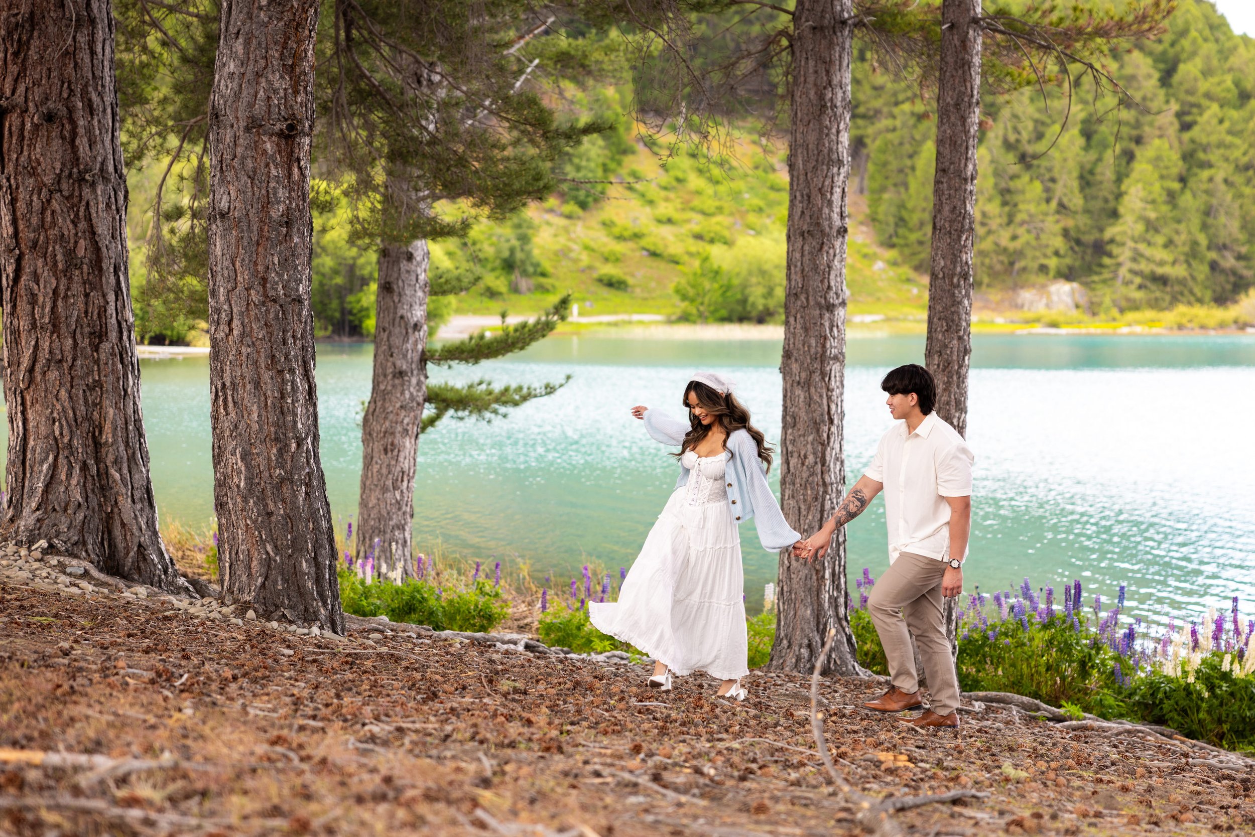 A couple walking hand in hand near a lake surrounded by trees, with the woman in a white dress and the man in a white shirt and beige pants.