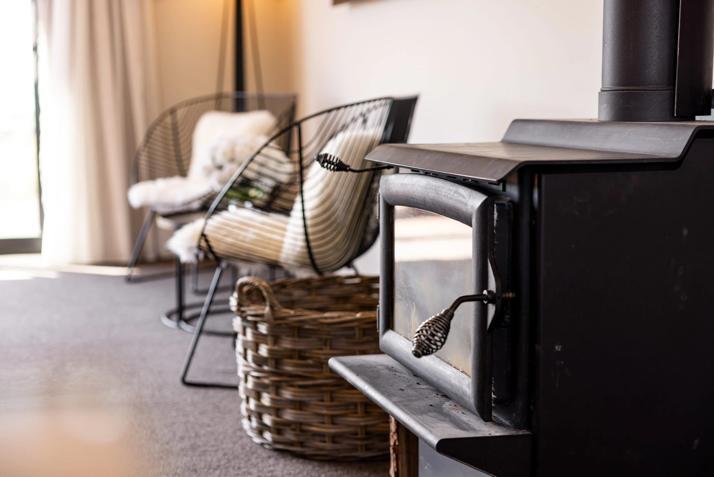 Living room with contemporary furniture, including a black wire chair with a white cushion, a woven basket, and a wood stove near a window with curtains.