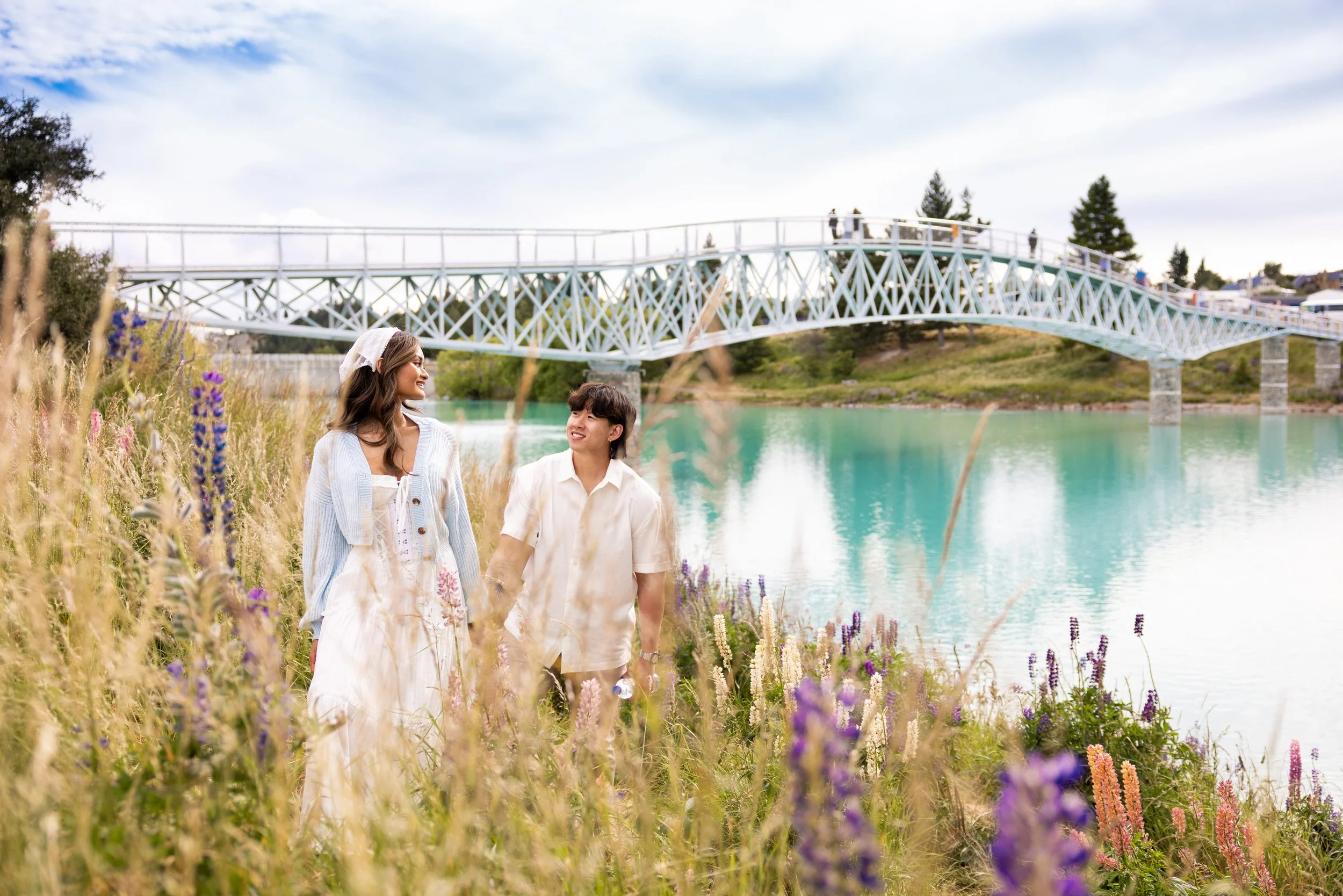 A young couple walking through a field of wildflowers near a body of water, with a bridge in the background and a partly cloudy sky.
