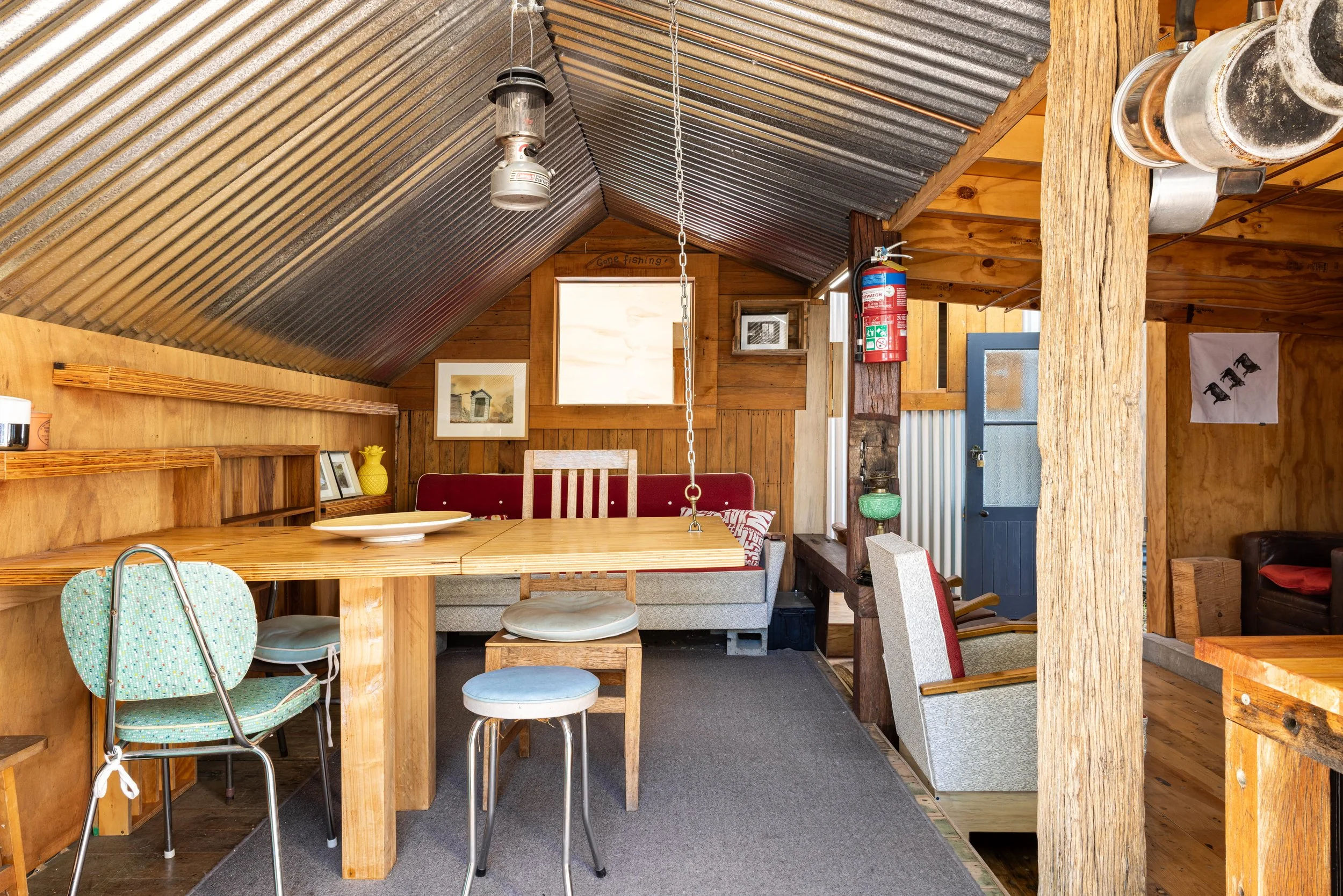 Interior of a cozy wooden cabin with a sloped, corrugated metal ceiling, featuring a dining area with mixed seating, a small window, and rustic decor.