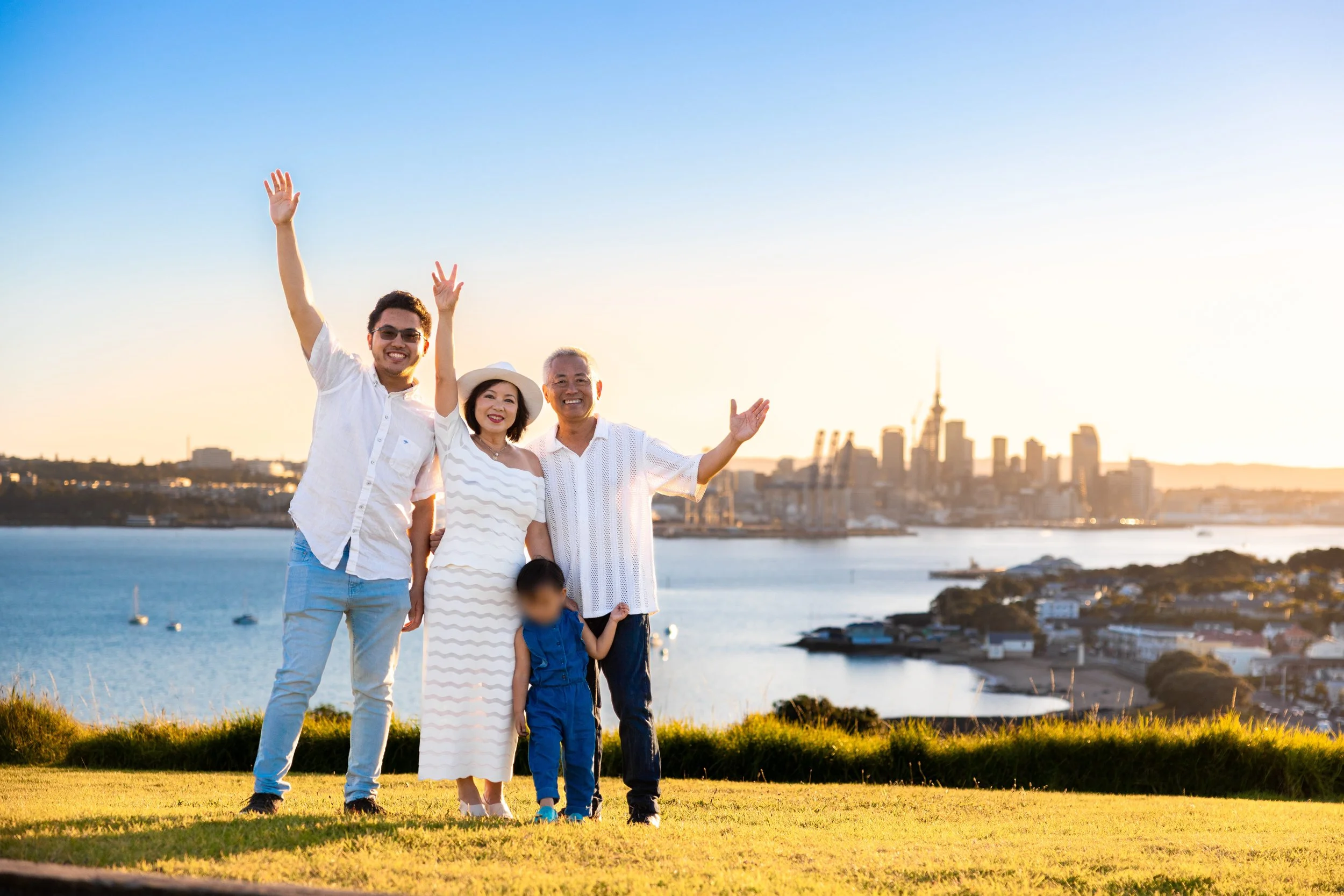 Family of four smiling and waving on a grassy hill with city skyline and water in the background during sunset.