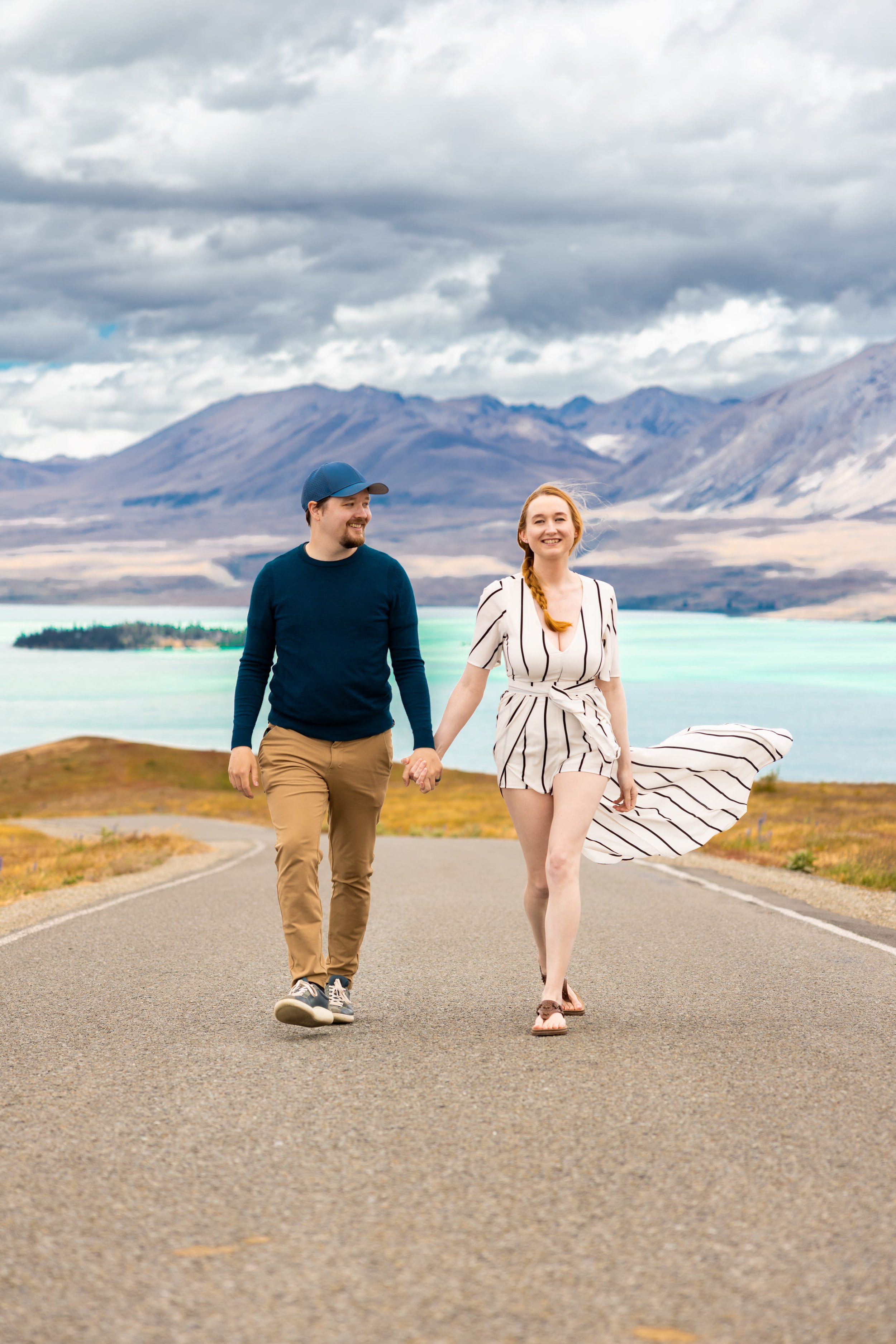 A couple walking hand in hand on a paved road in a scenic landscape with mountains, a lake, and cloudy skies in the background.