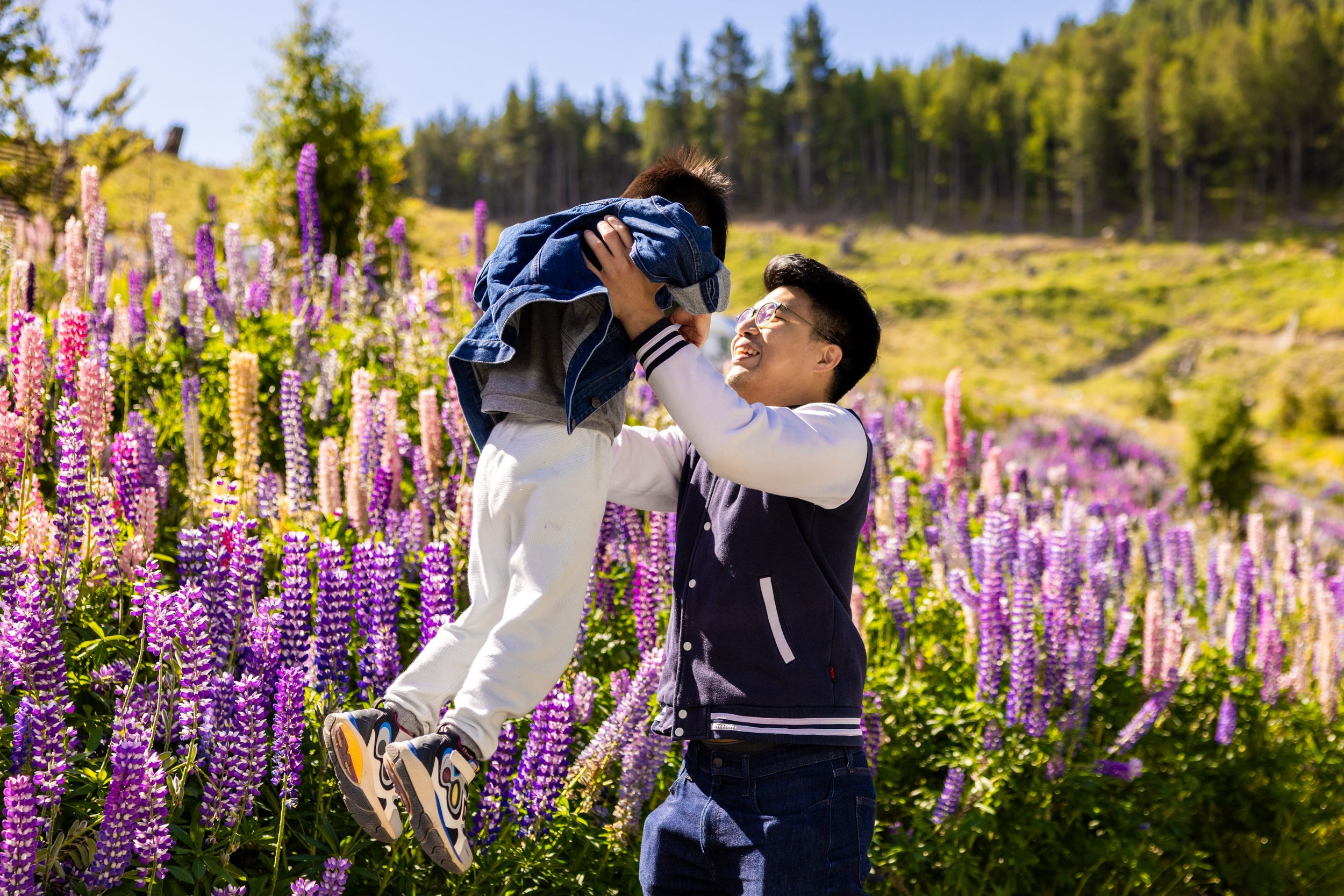 A man lifting a child in a field of purple and pink flowers with trees in the background on a sunny day.
