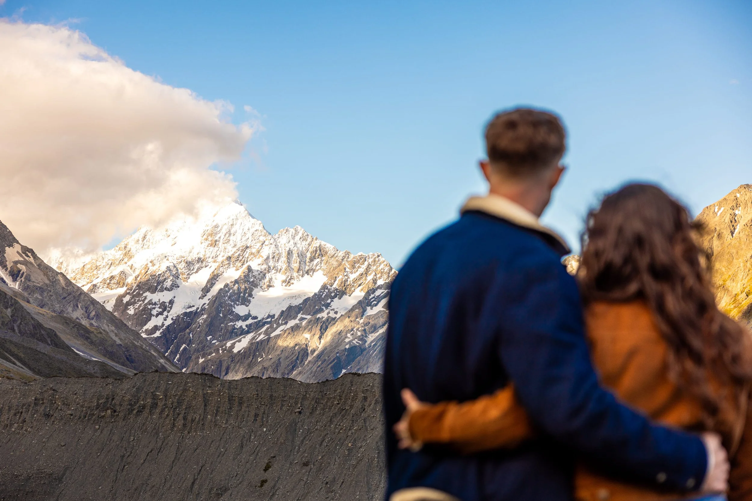 A romantic couple standing close together, facing a mountain range with snow-capped peaks and a partly cloudy sky.