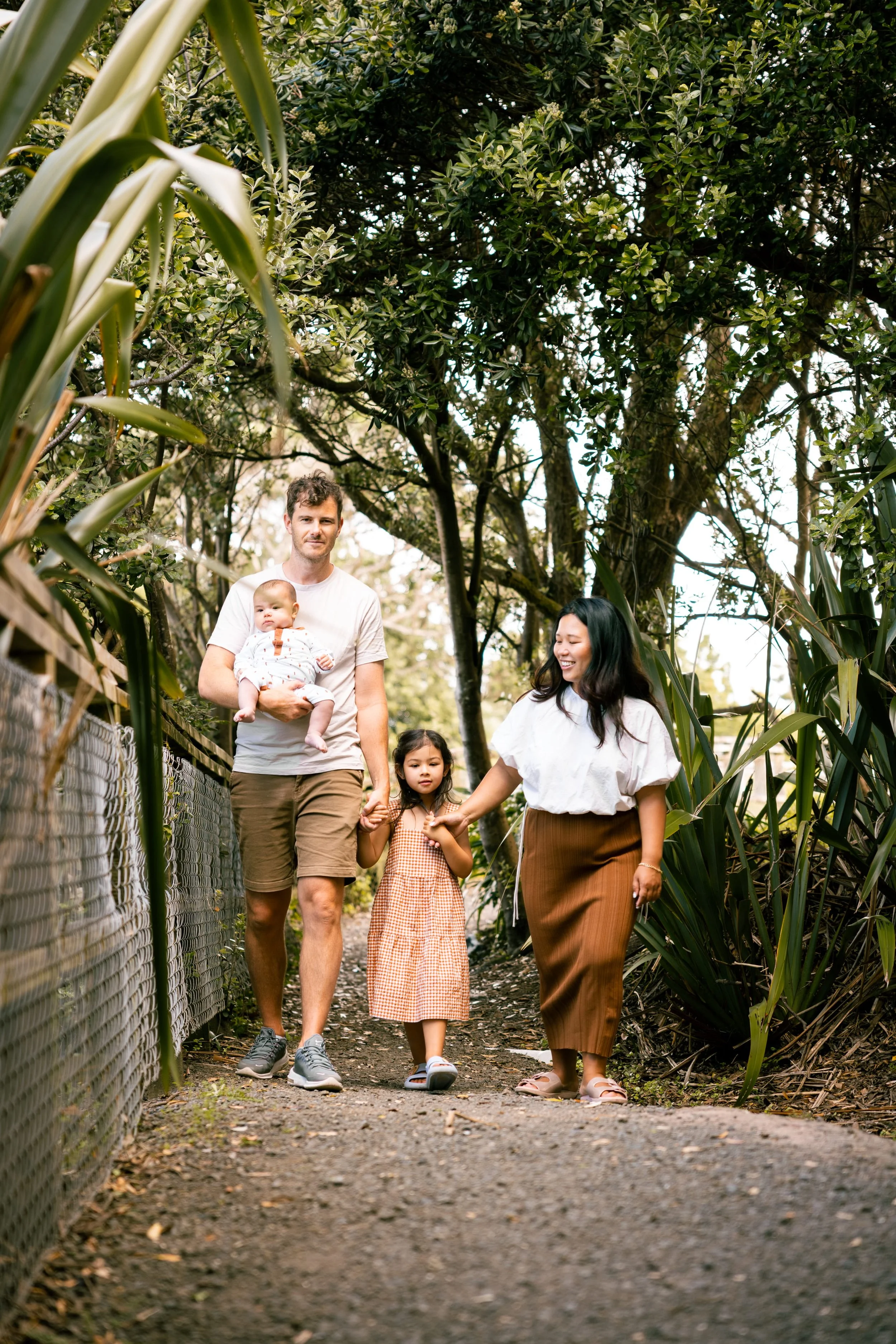 Family walking outdoors on a dirt path surrounded by trees and greenery, holding hands and smiling.