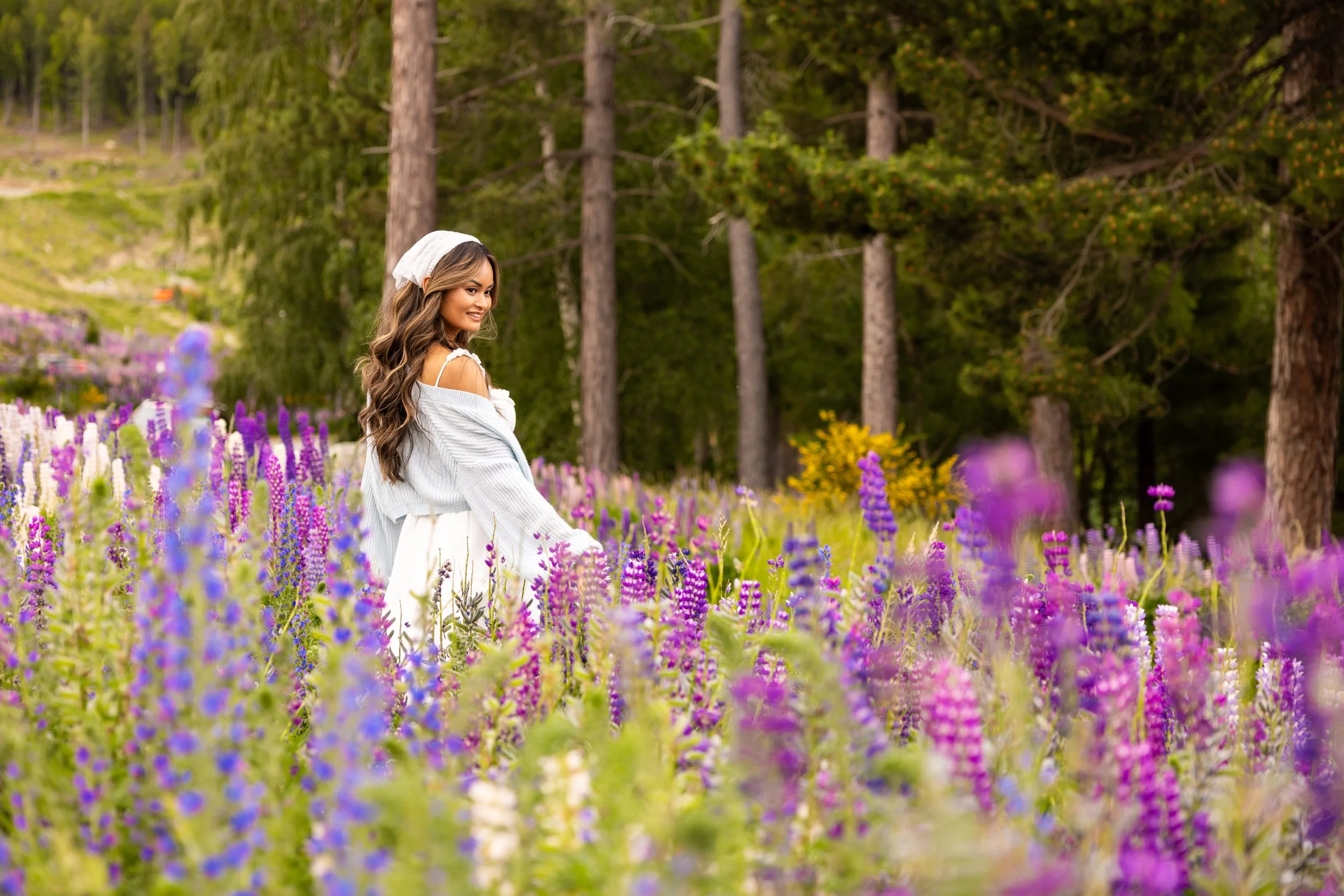 A young woman with long wavy brown hair, wearing a white off-shoulder top and a white skirt, stands in a colorful flower field with purple, pink, and yellow flowers, and tall trees in the background.