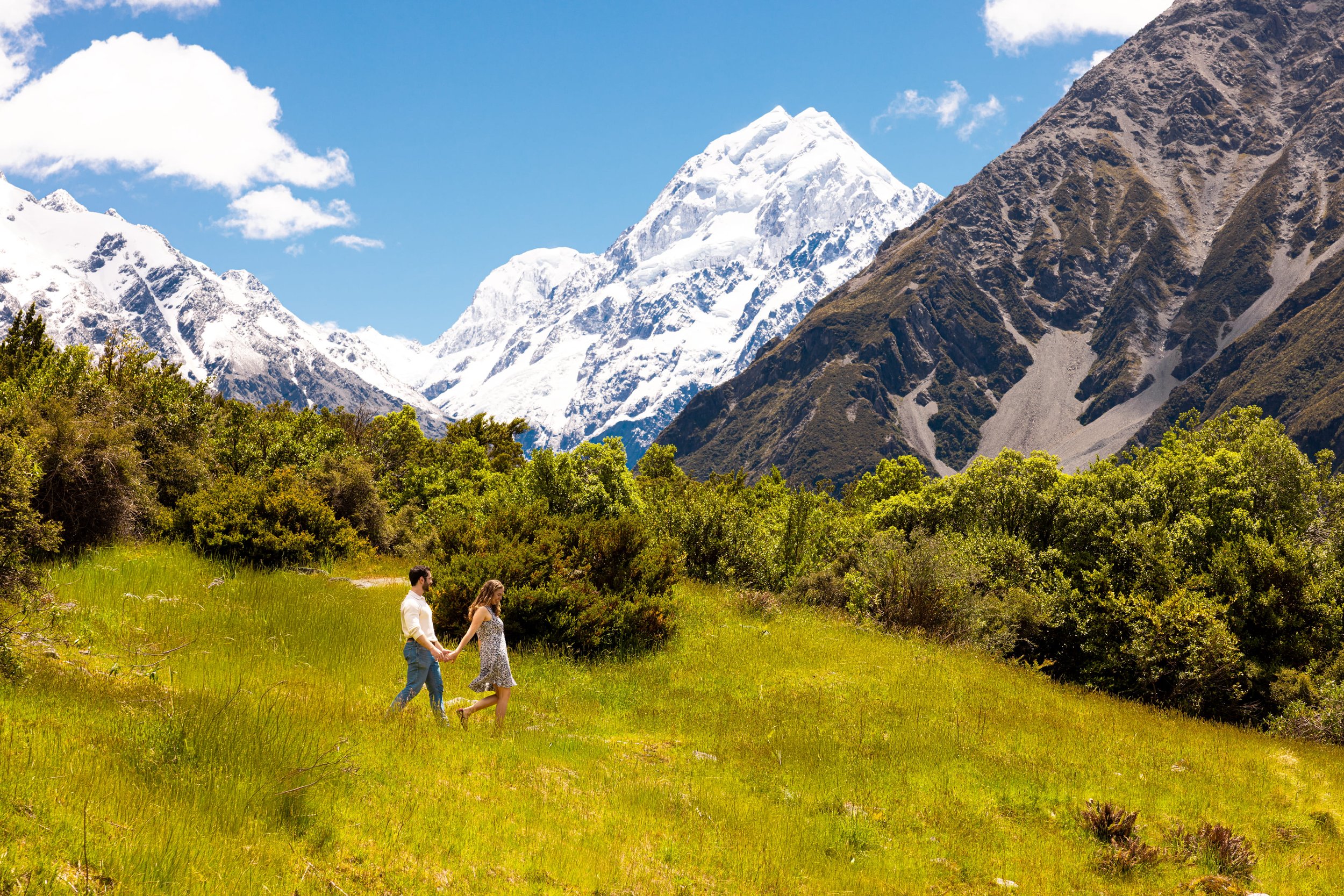 A man and woman holding hands walking through a grassy meadow with green bushes, snow-capped mountains in the background, under a partly cloudy blue sky.