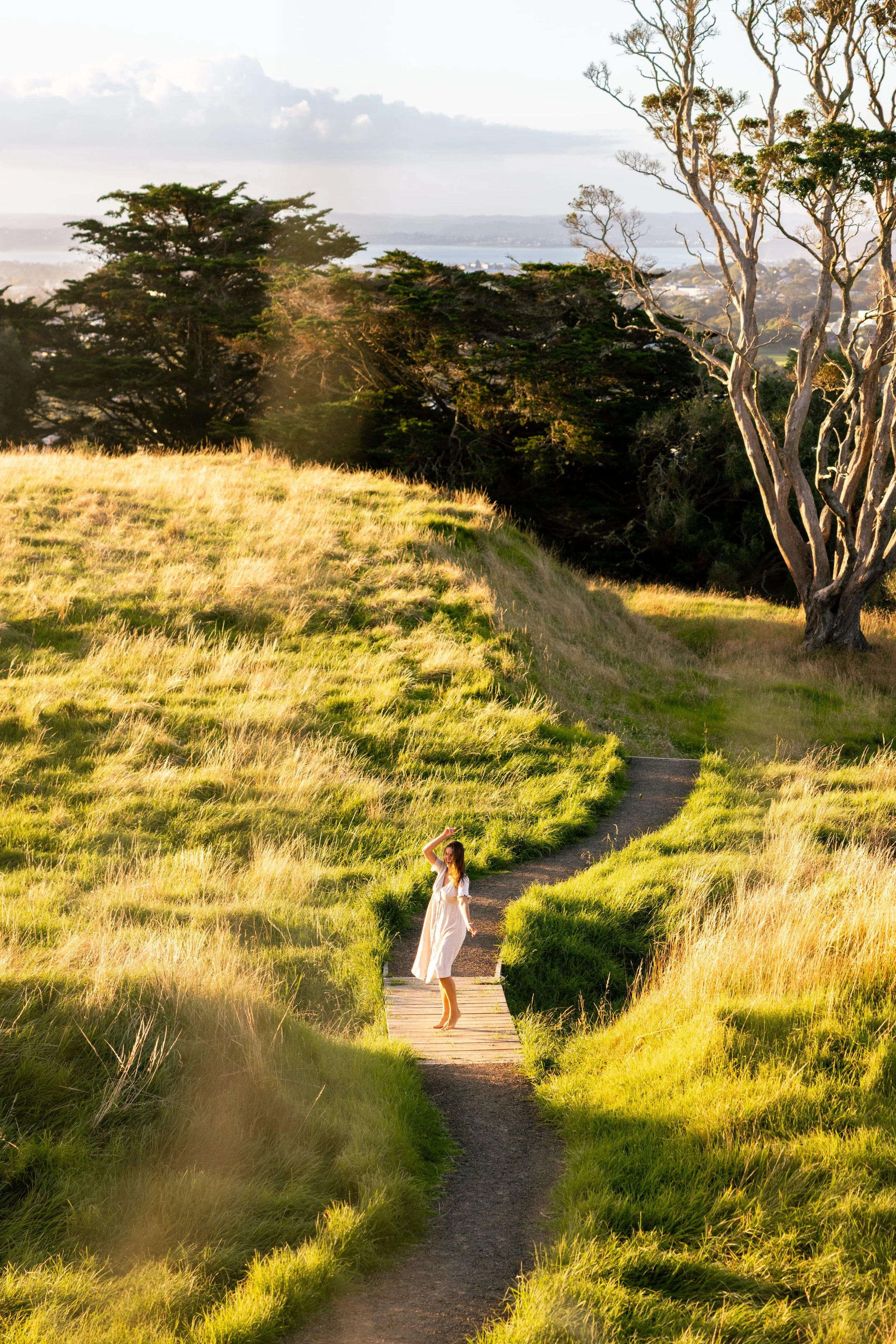 A woman in a white dress standing on a narrow winding wooden bridge in a grassy field during daylight with trees and a distant city in the background.