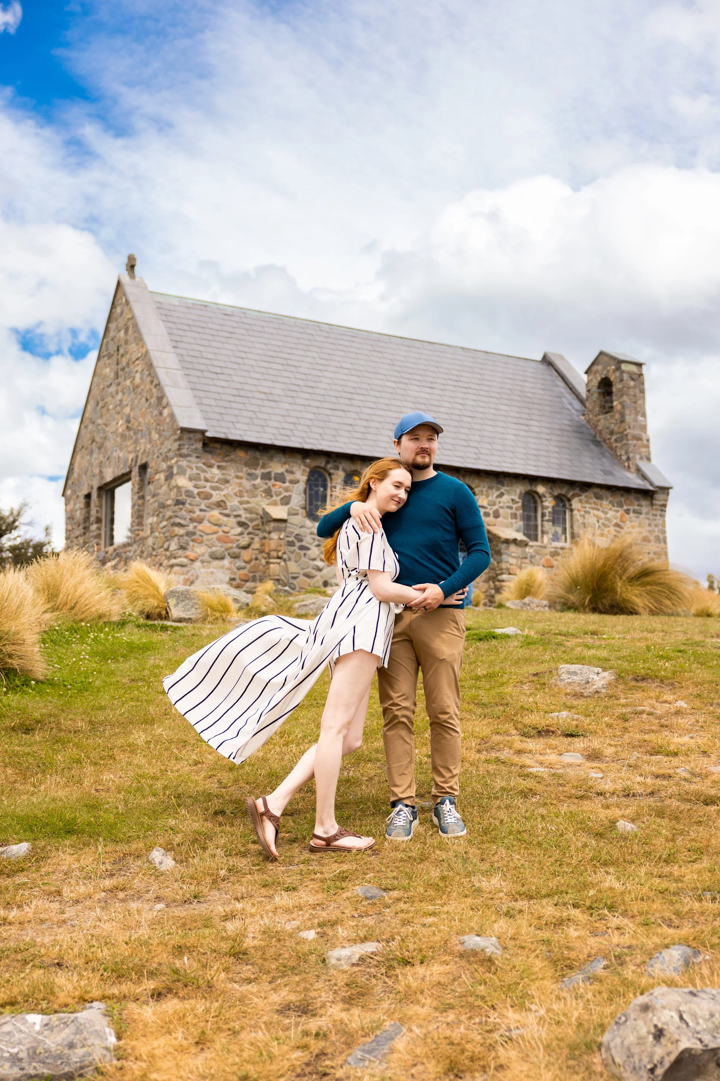 A couple standing in front of a stone church, embracing and smiling, the woman wearing a white dress with navy stripes and the man in a blue shirt and khaki pants.