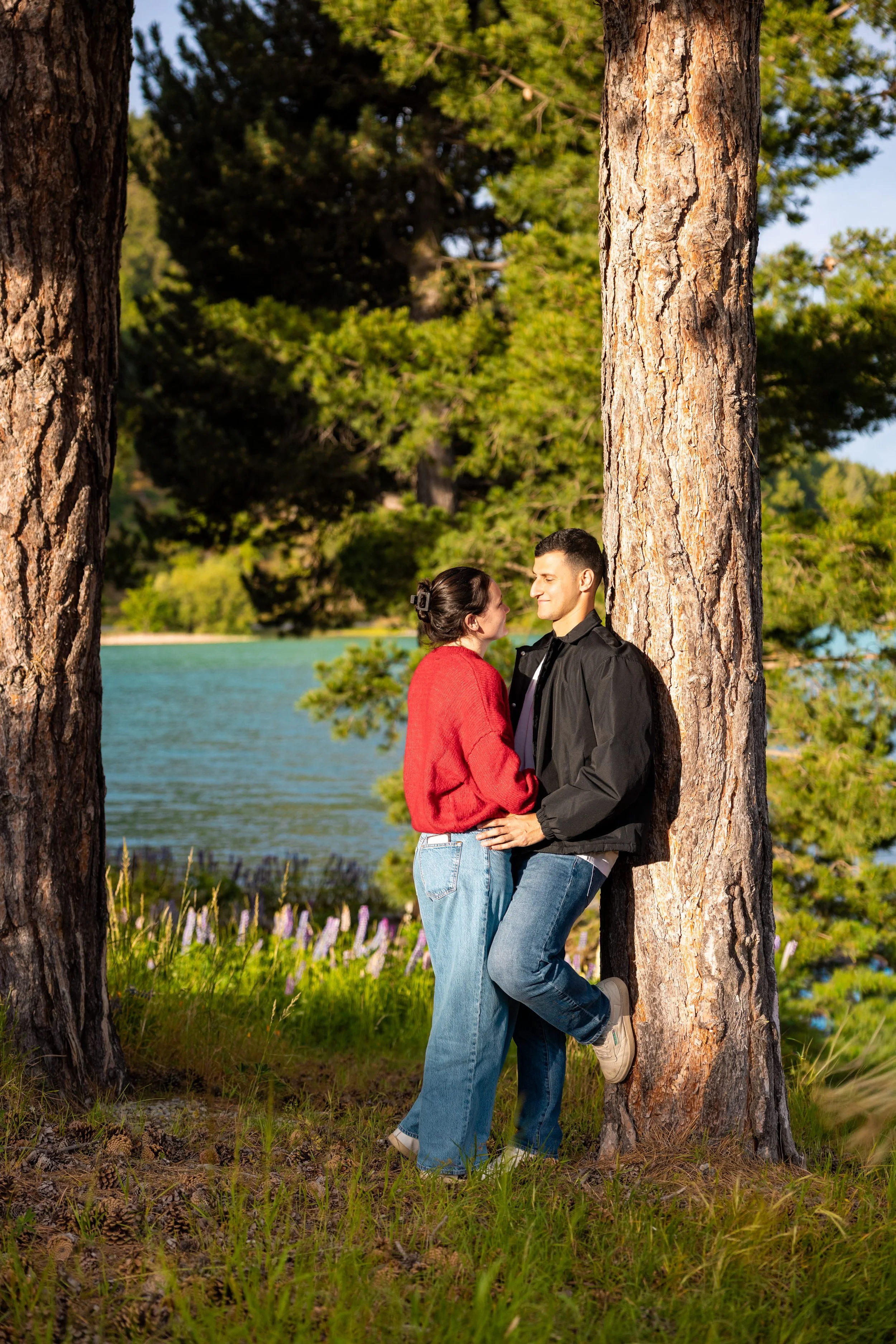 A couple standing near two large pine trees by a lake, facing each other, with the woman wearing a red sweater and the man in a black jacket, looking at each other lovingly.