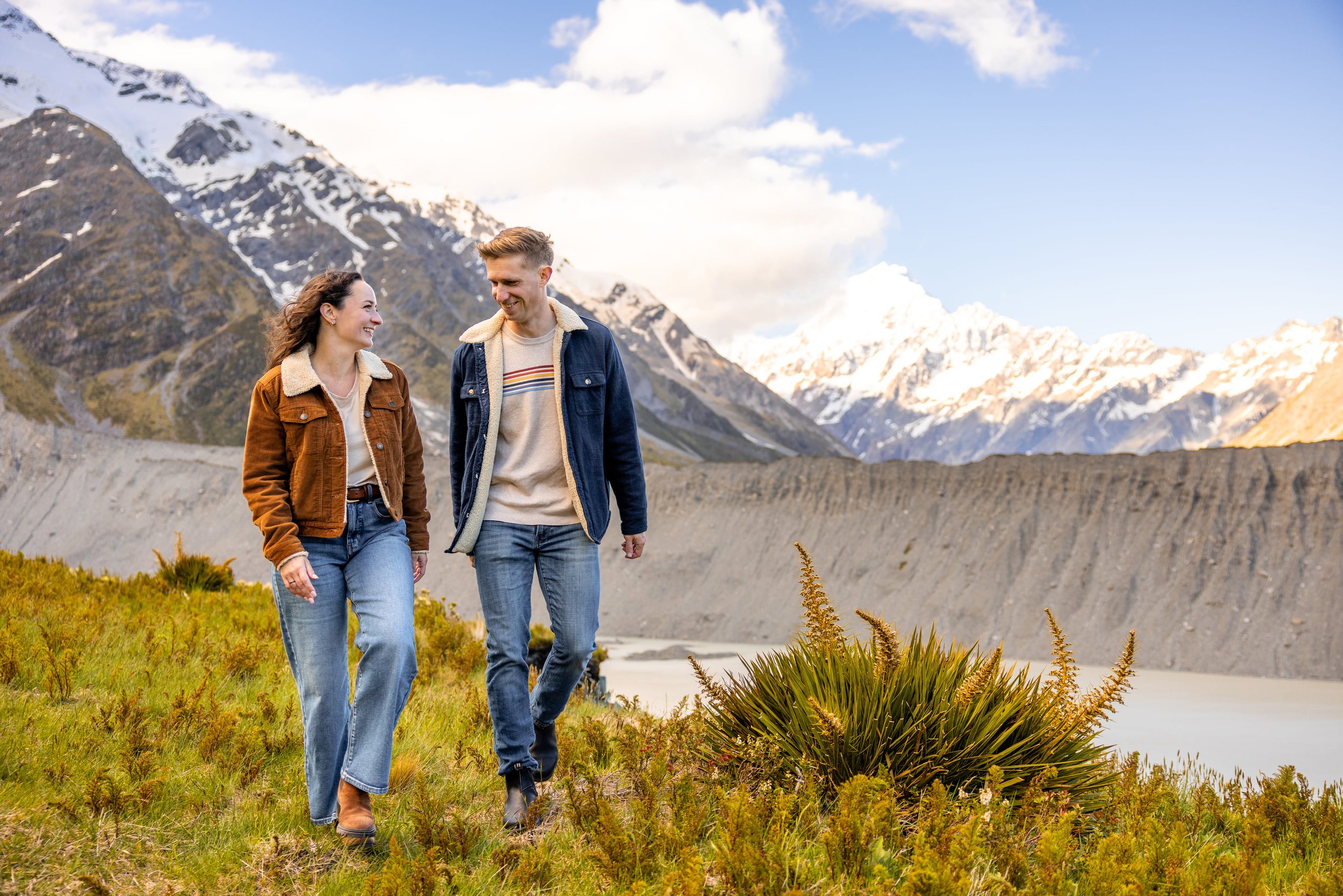 A man and woman walking together outdoors in a scenic mountain landscape with snow-capped peaks and a lake in the background, smiling and enjoying each other's company.