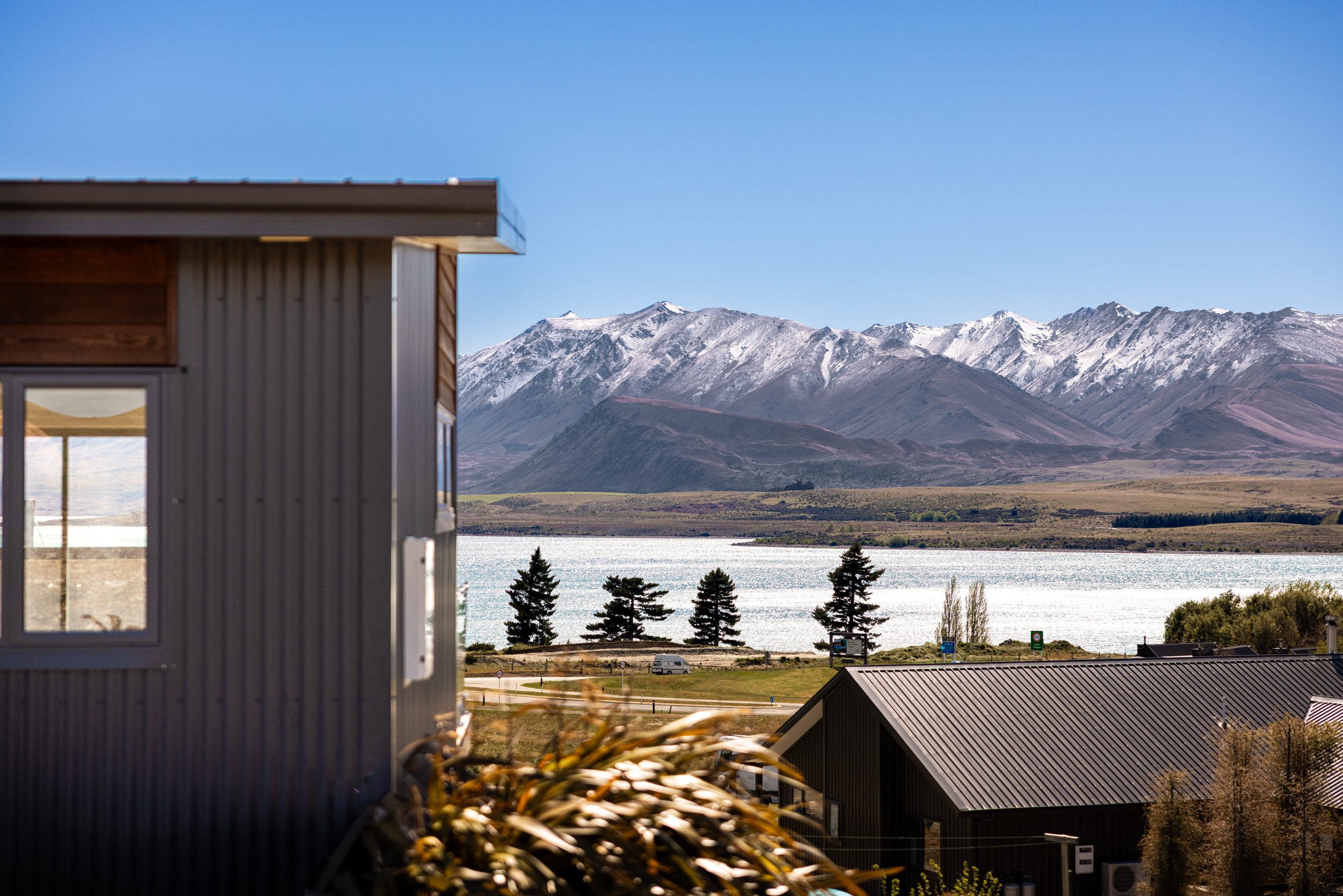 A scenic view of snow-capped mountains over a body of water with houses and trees in the foreground.