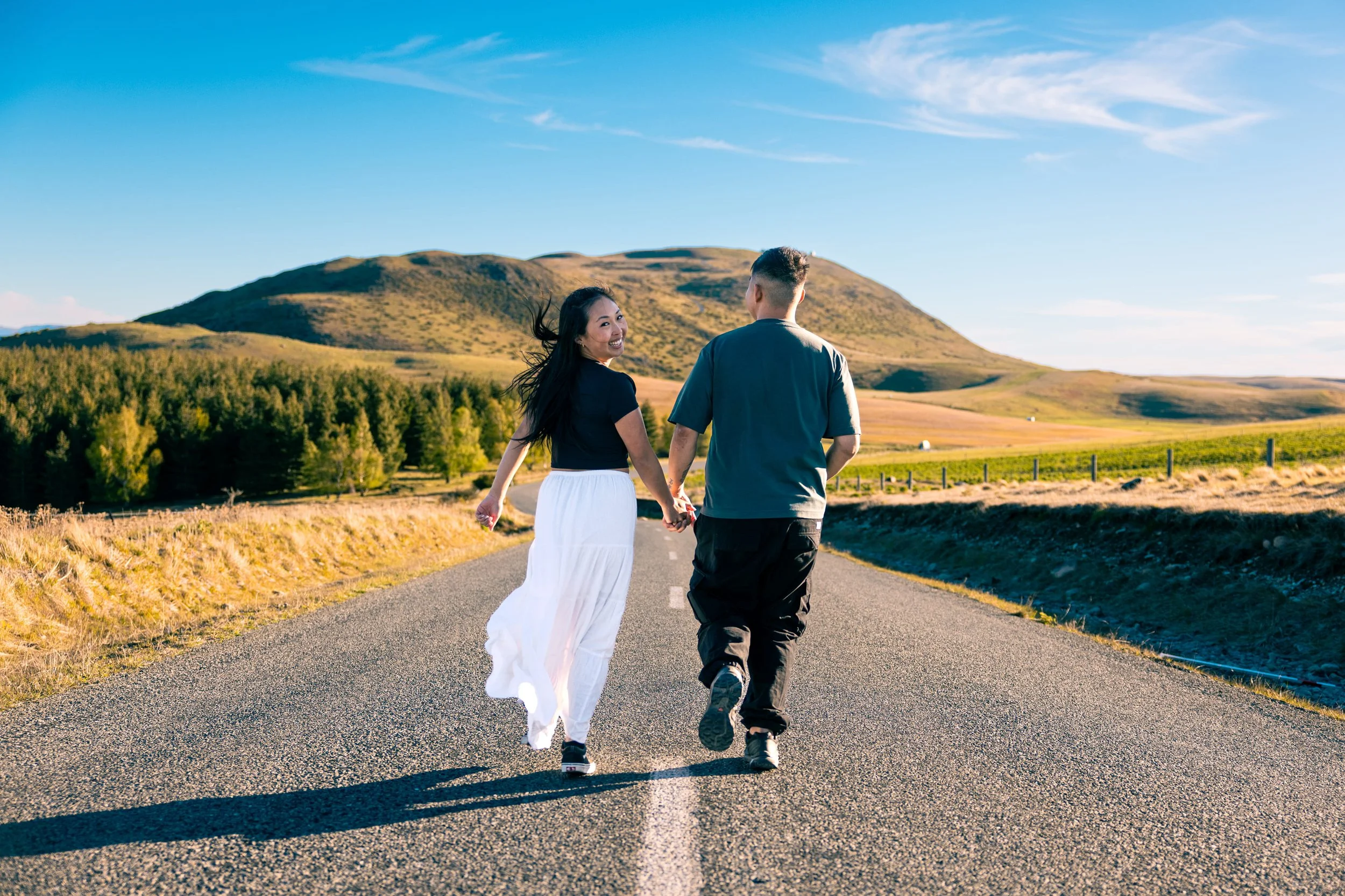 Couple walking hand-in-hand on an open road in a rural landscape with hills and trees, under a partly cloudy sky.