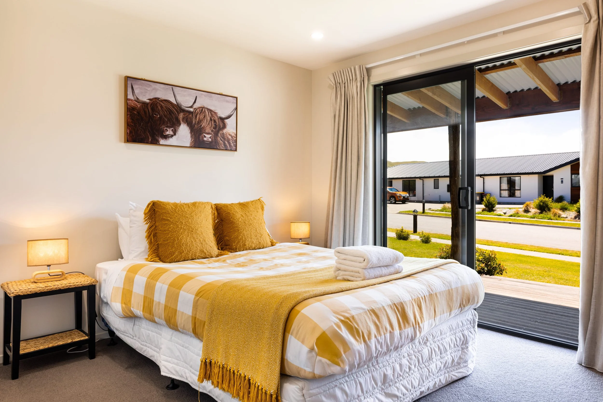 Bedroom with a bed covered in yellow and white checkered bedding, two brown pillows, and folded white towels, next to a sliding glass door showing a view of houses and greenery outside.