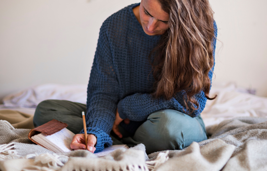 Person sitting on a bed writing in a notebook with a pencil, wearing a blue sweater and green pants.