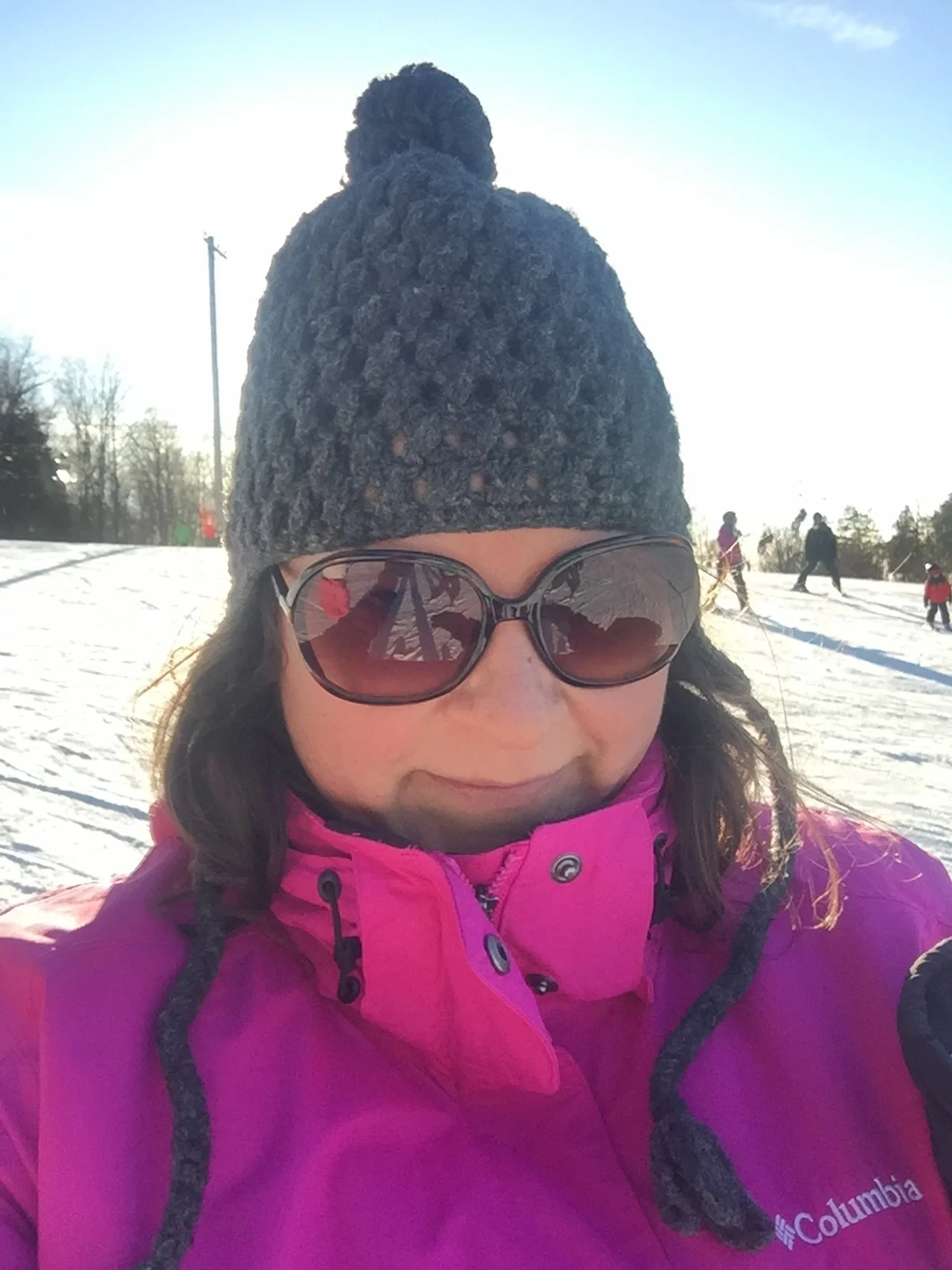 Amara dressed in a pink Columbia jacket and wearing large sunglasses, a gray knit hat outdoors on a snowy day with other people skiing in the background.