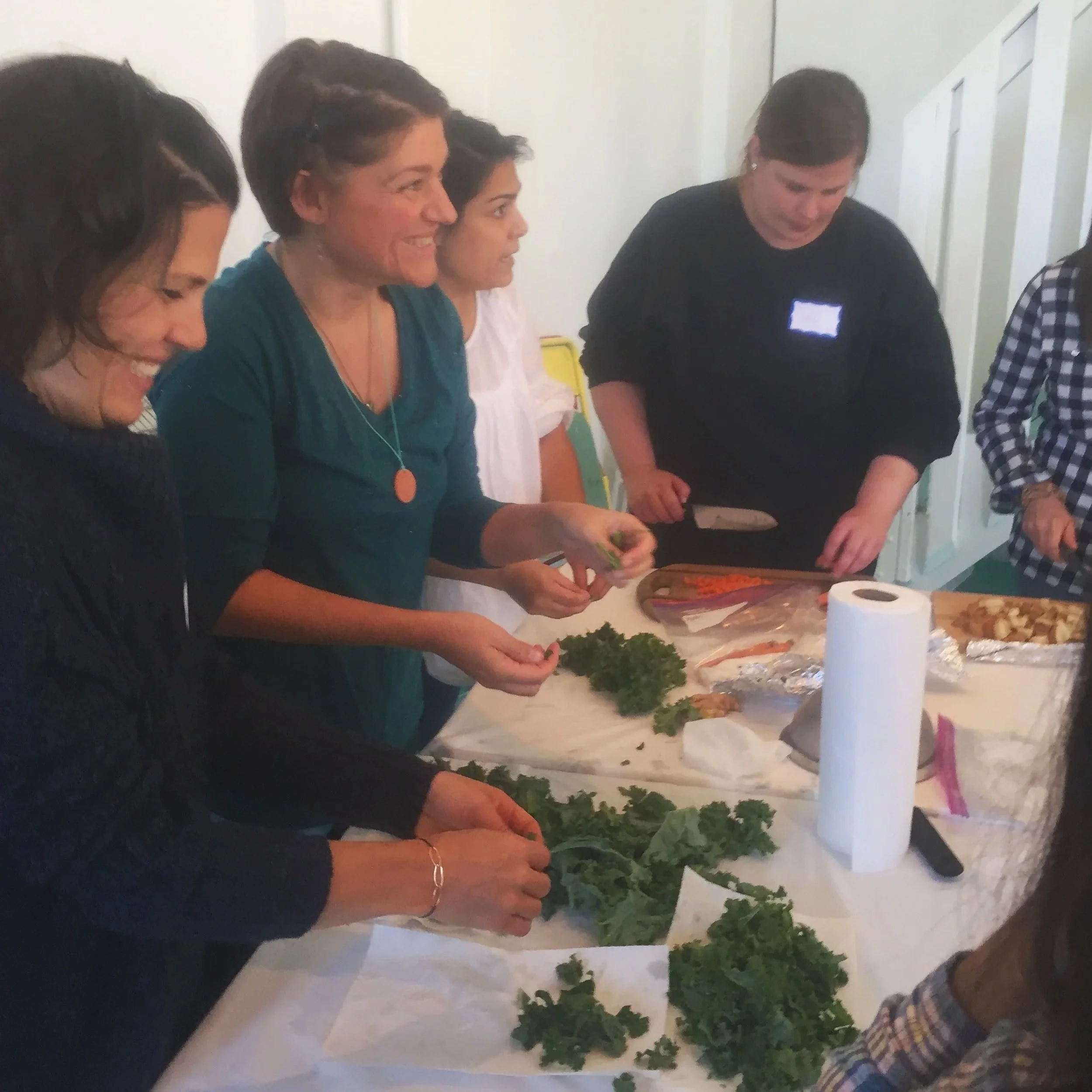 Group of mothers in a cooking class together at a table with herbs and vegetables, in a kitchen setting.