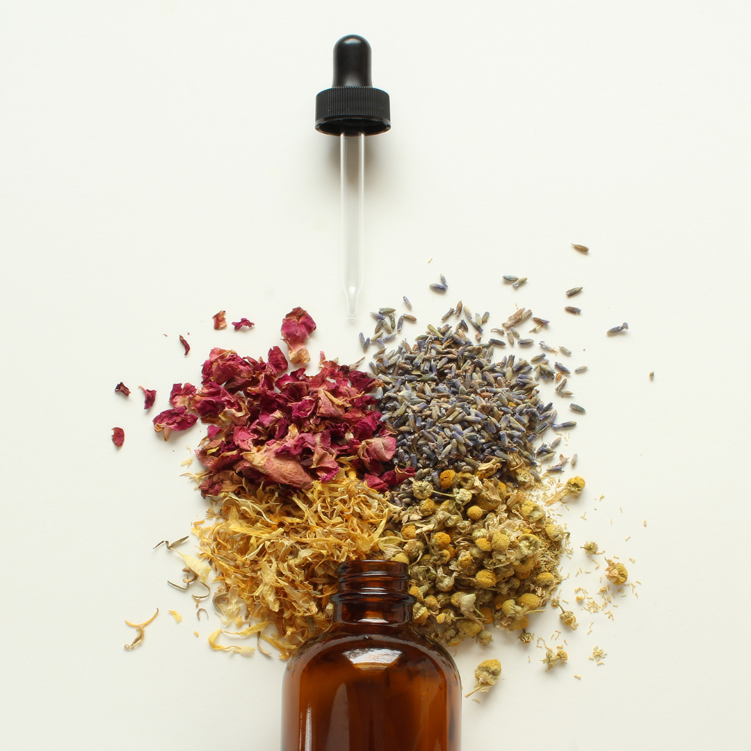 A brown glass dropper bottle with dried herbs, including red rose petals, lavender, chamomile, and calendula, arranged in a circle around it on a white background.