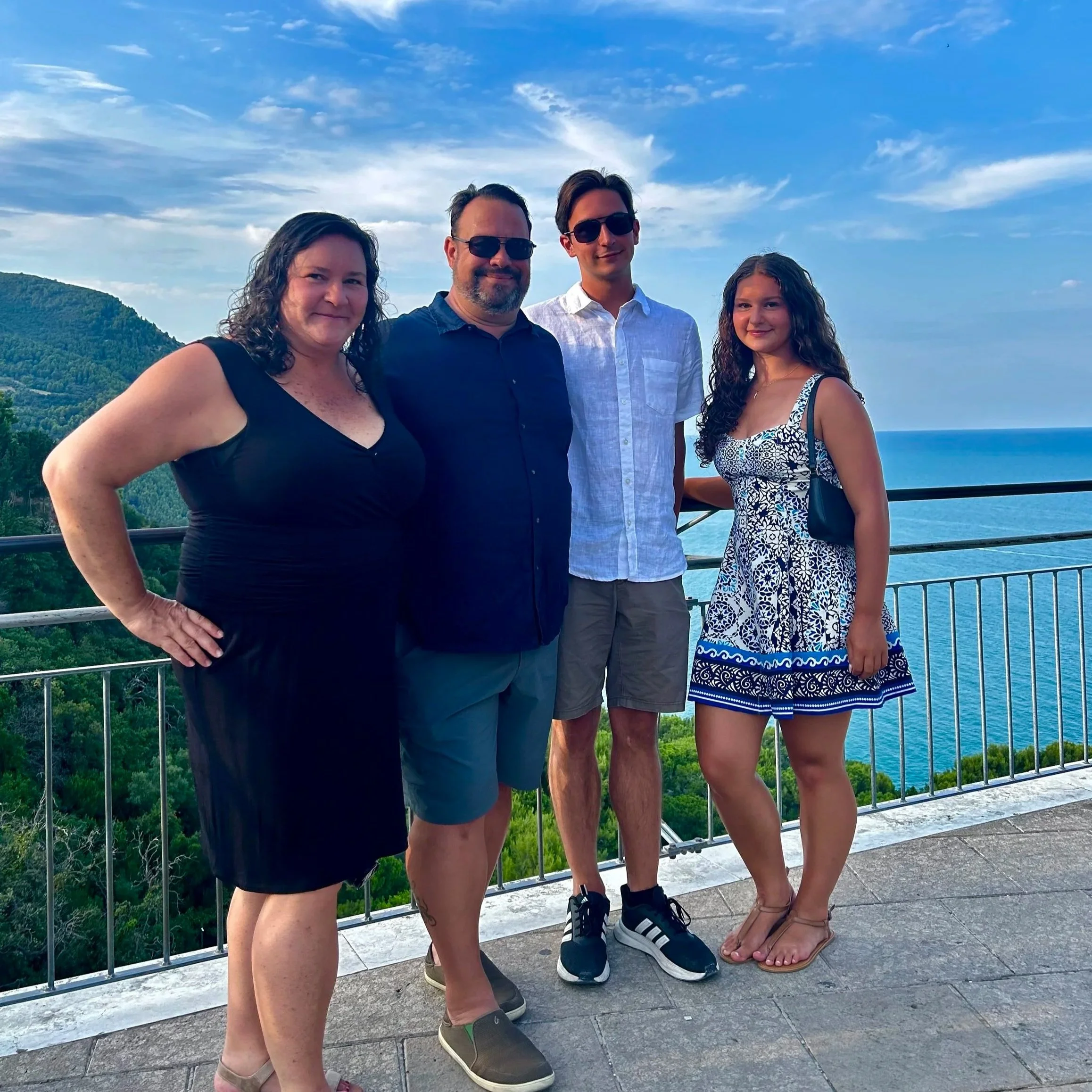 Four people standing on a balcony with scenic view of mountains, greenery, and water in the background on a cloudy day.