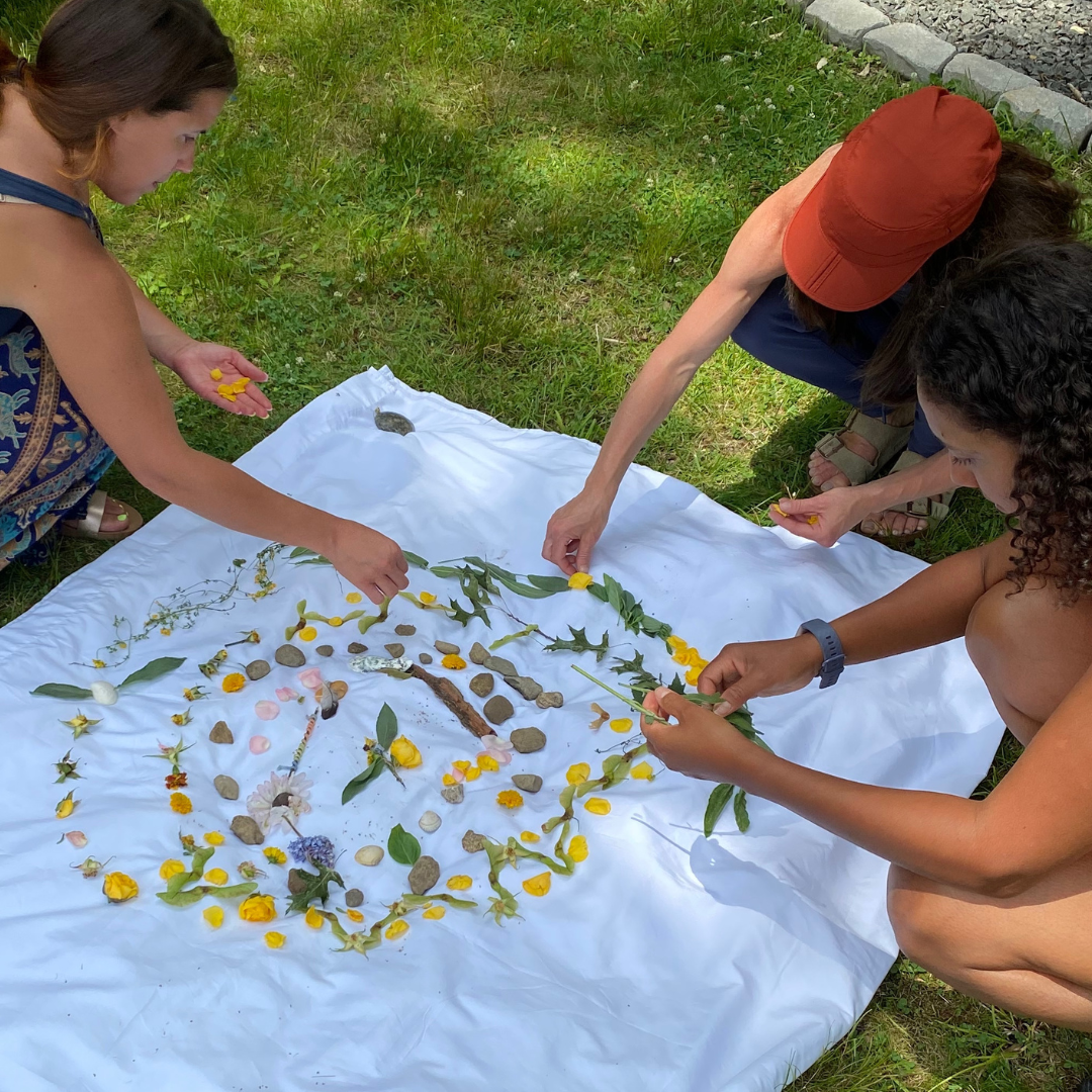 Three people arranging yellow petals, leaves, and stones in a circular pattern on a white cloth outdoors.