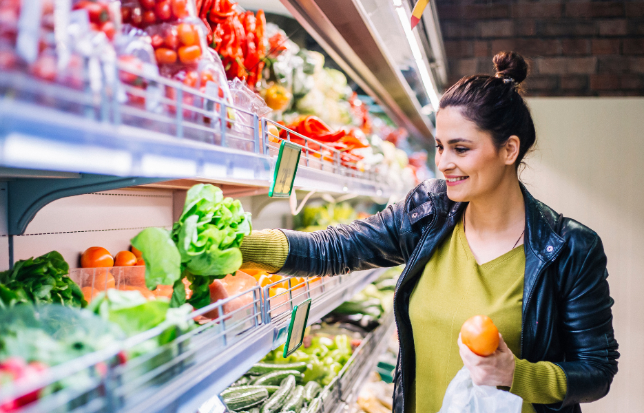 Woman shopping for fresh vegetables in a grocery store, holding a tomato and reaching for lettuce on a shelf filled with various produce items.