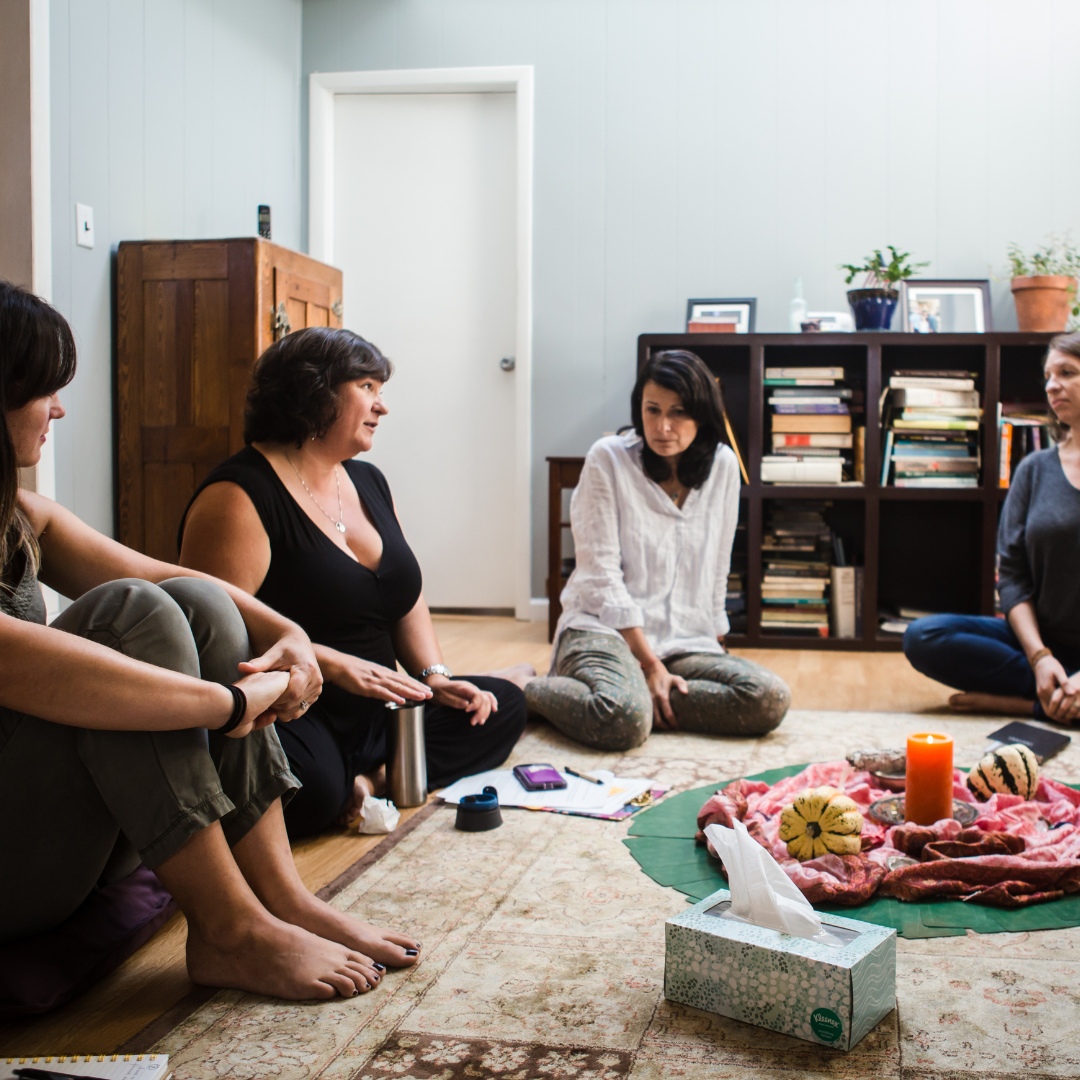 A group of women sitting on the floor in a circle, engaged in a discussion or support group setting. The room features bookshelves in the background, a rug, and a centerpiece with a candle and decorative items. A box of tissues is placed nearby.