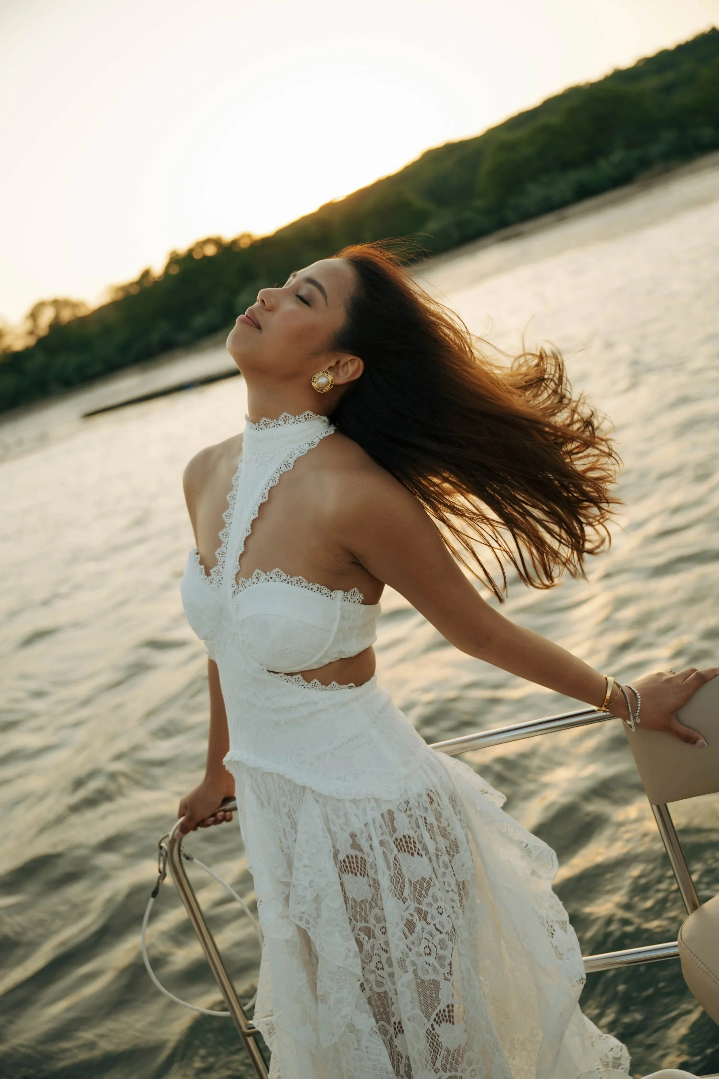 A woman in a white lace dress enjoying the sunset on a boat, with her eyes closed and hair flowing in the wind, near a green hilly landscape.