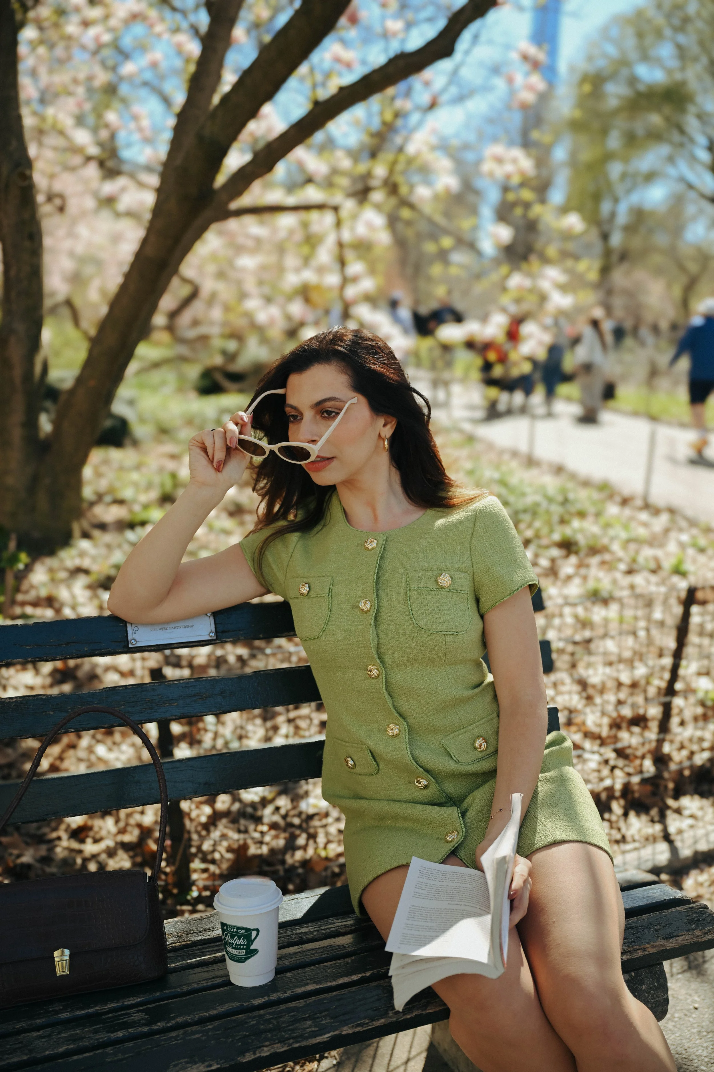 Woman sitting on a park bench, wearing a green dress, holding sunglasses, with a magazine in her lap, in a park with blooming trees, coffee cup on the bench, and other people in the background.