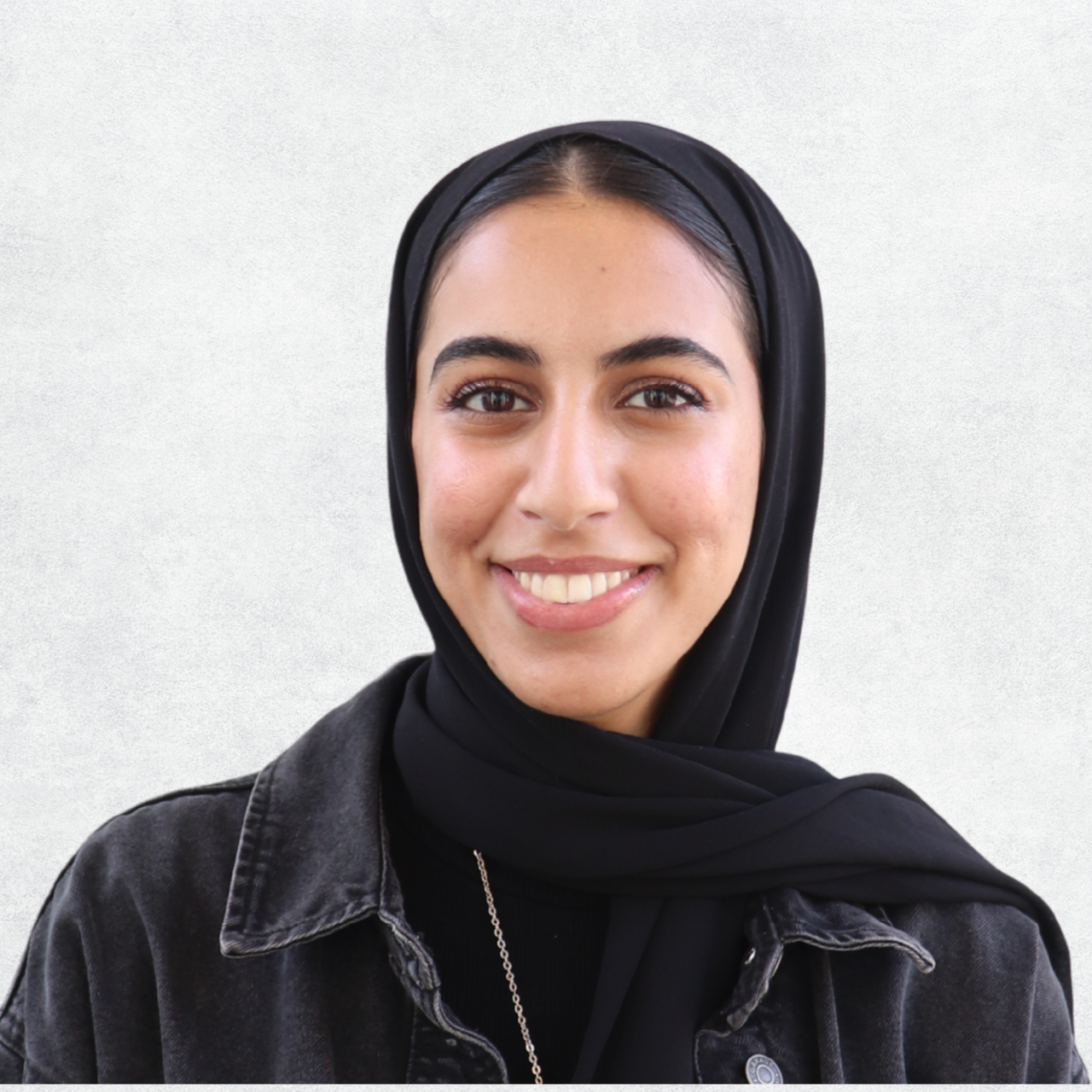 A young woman with light skin wearing a beige hijab and earrings, smiling at the camera.
